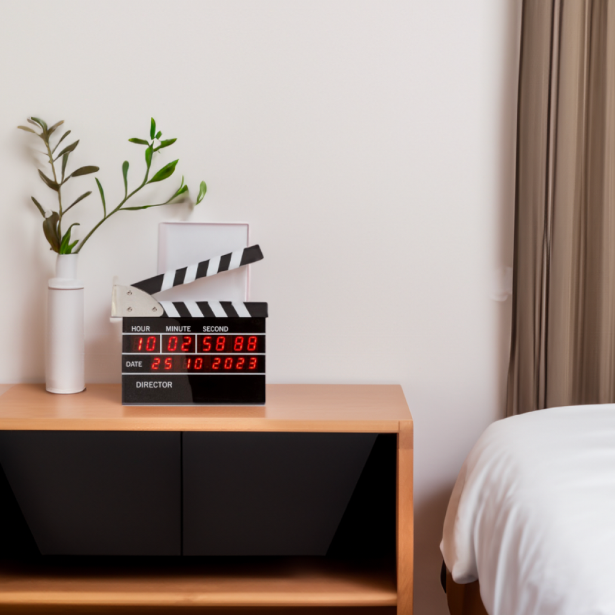 A digital film clapperboard timer with red numbers sits on a bedside table near a vase.