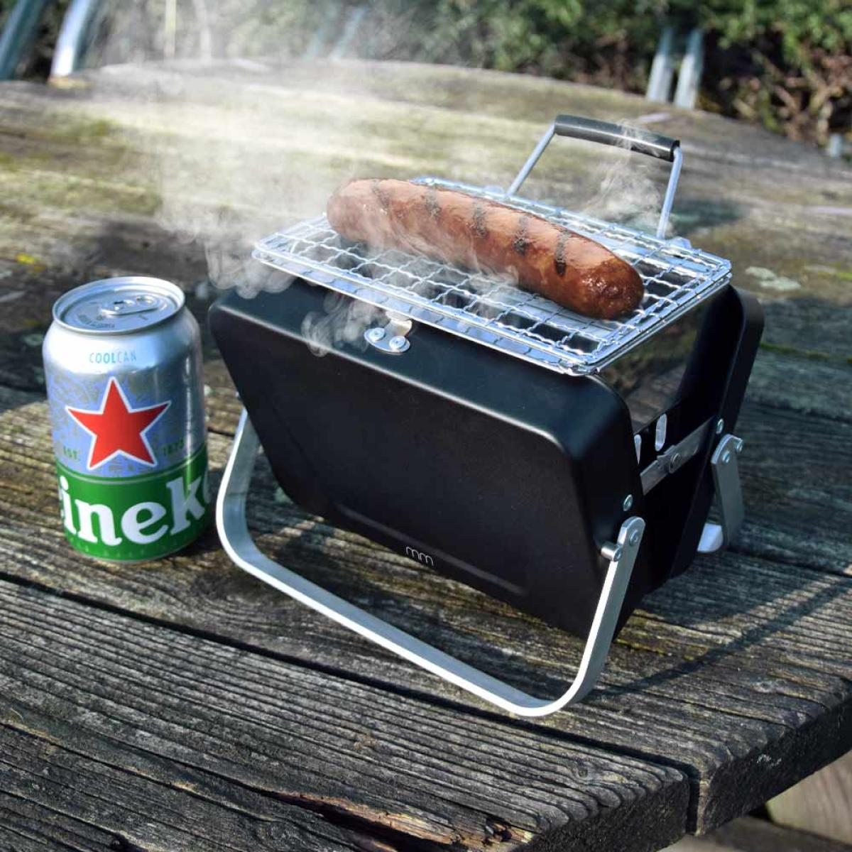 A sausage is grilling on a small barbecue, next to a can of beer.