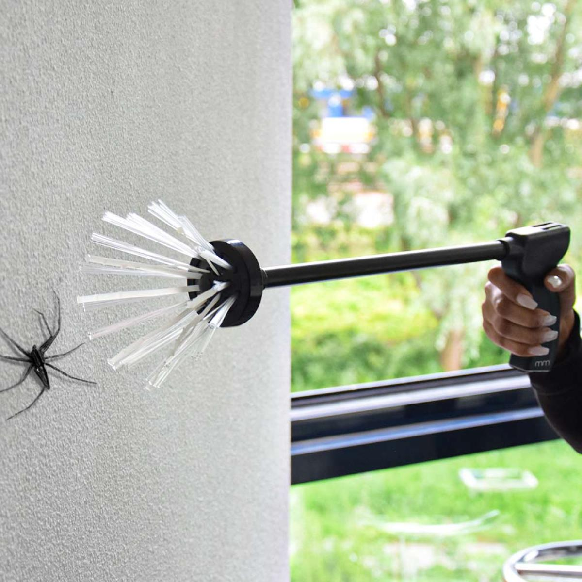 Een spinnenvanger met borstels vangt een spin op een muur naast een raam.