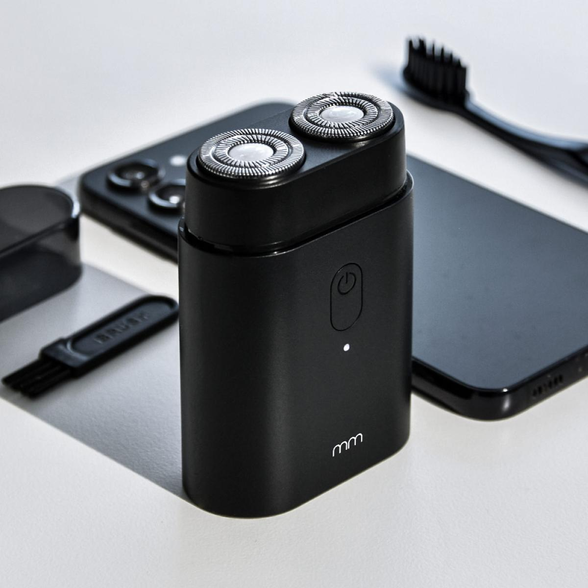 A black electric shaver with two heads, beside it a smartphone, a brush, and other accessories.