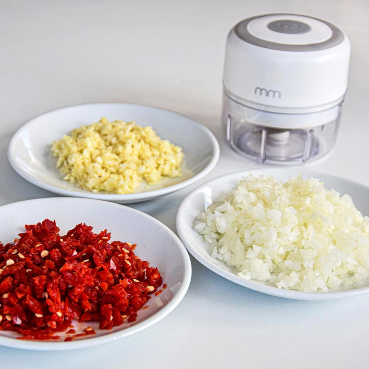 Chopped chilies, garlic, and onions next to a small white mini chopper.