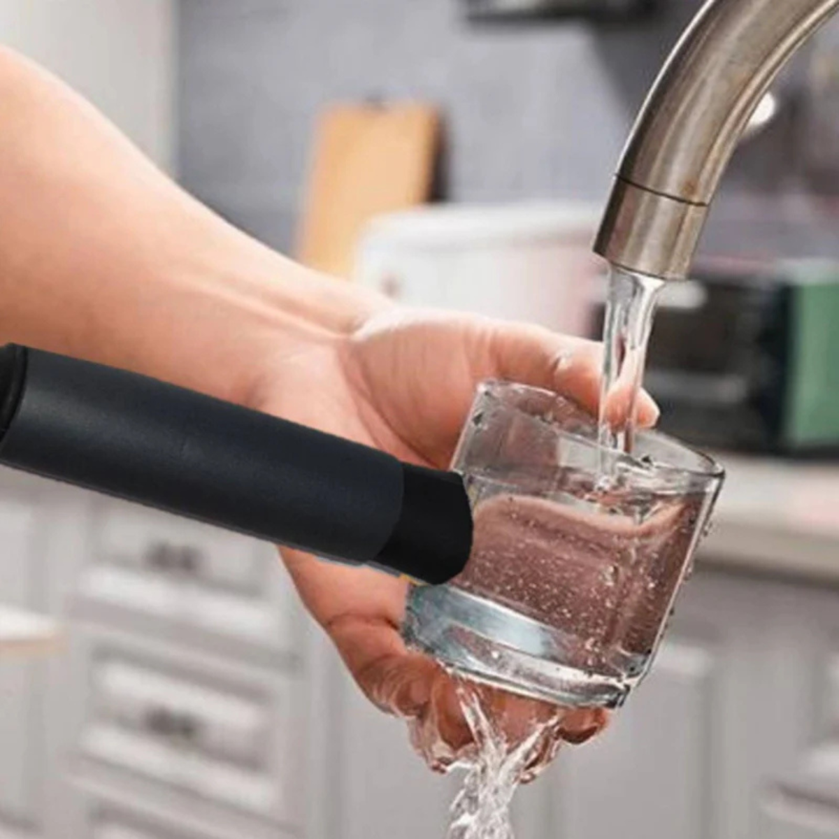 A hand holds a glass under running water. A black device is beside it.