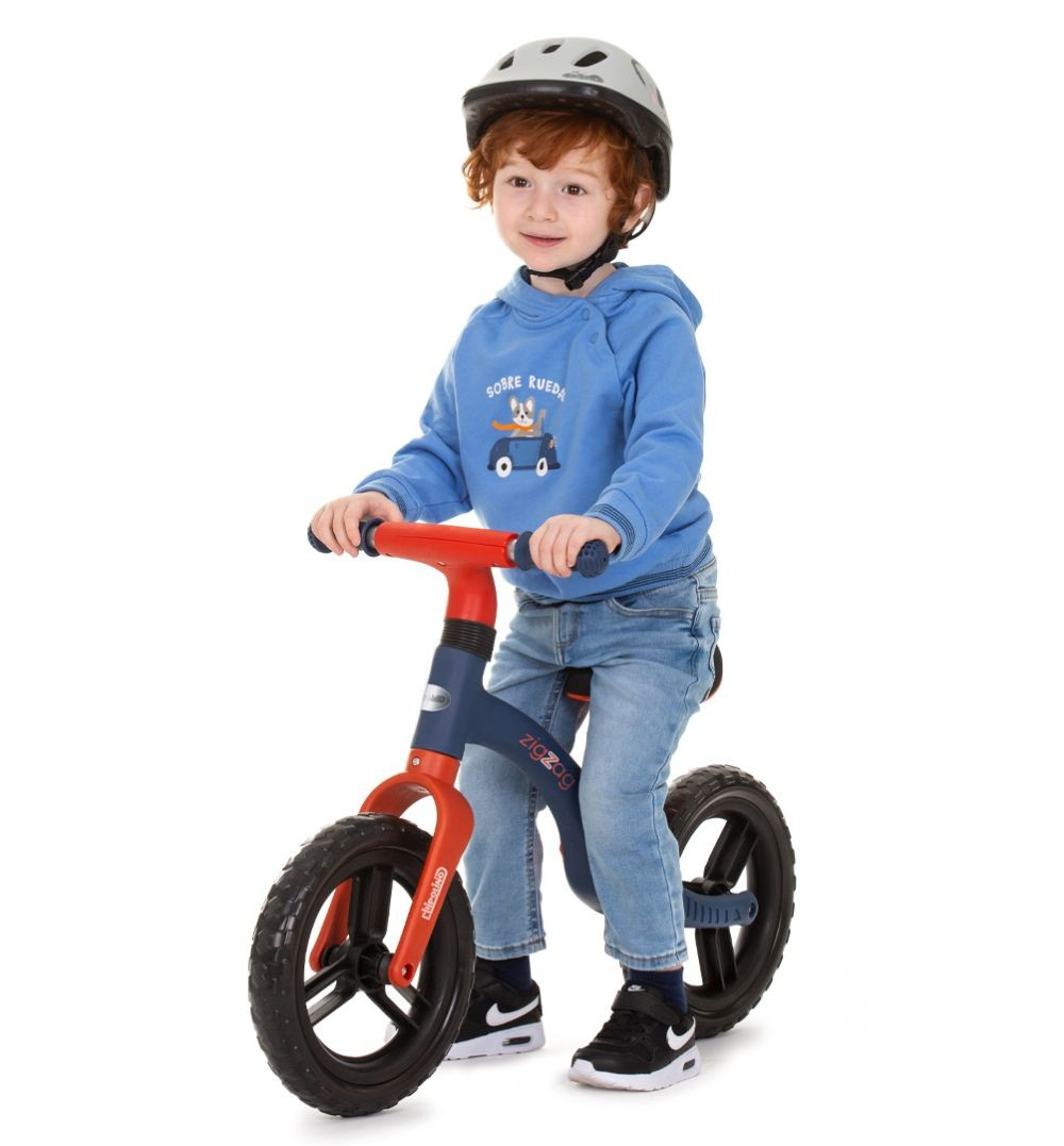 Ein kleiner Junge mit roten Haaren fährt ein blau-rotes Laufrad und trägt einen grauen Helm.