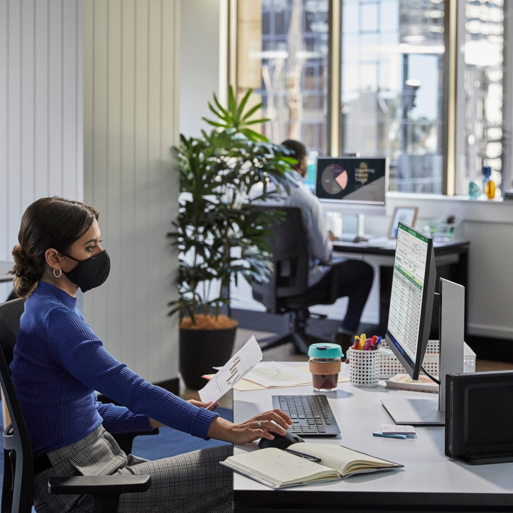 Kobieta w biurze w masce pracuje przy komputerze, kolega w tle.