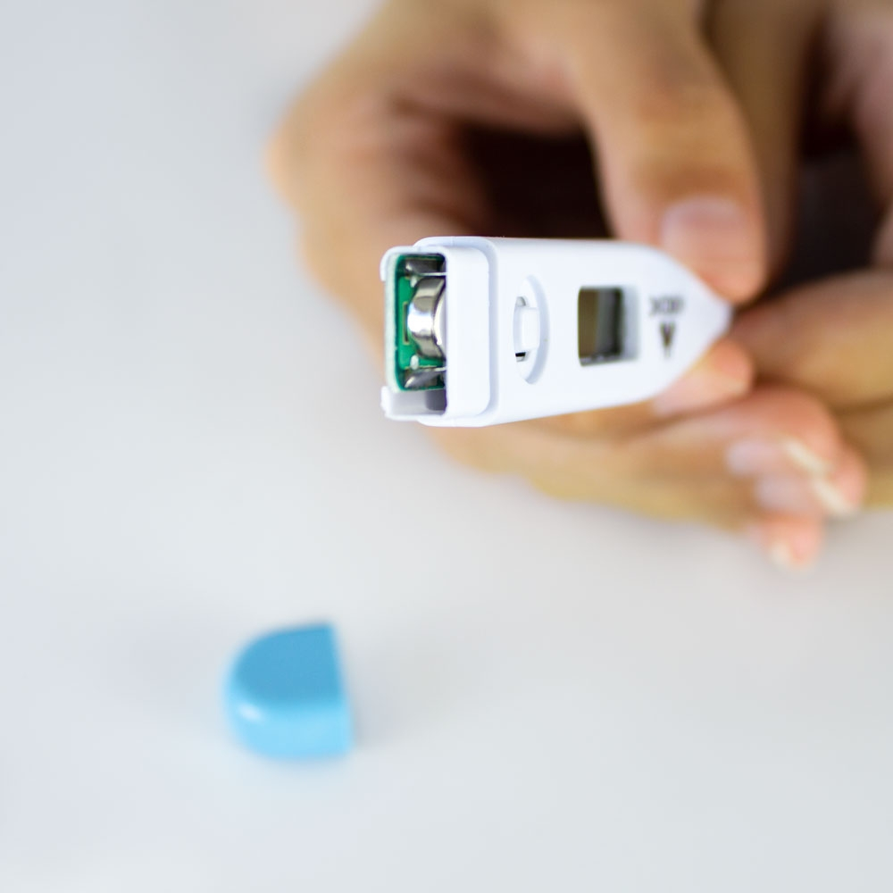 Una mano sostiene un termómetro digital blanco sin tapa de batería. Una tapa azul yace cerca.
