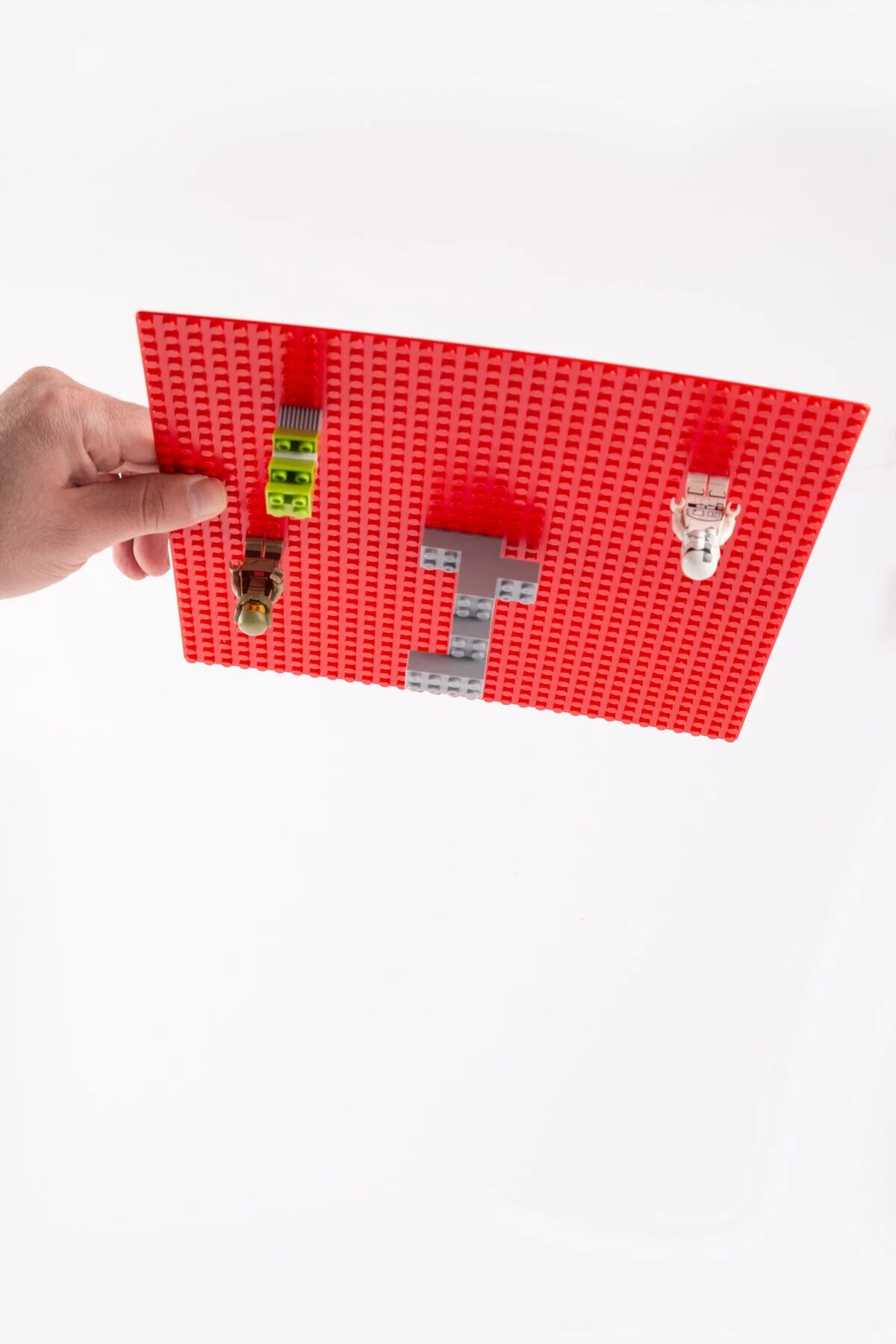 Bir el, küçük figürler ve gri tuğlalarla kırmızı Lego taban plakasını tutuyor.