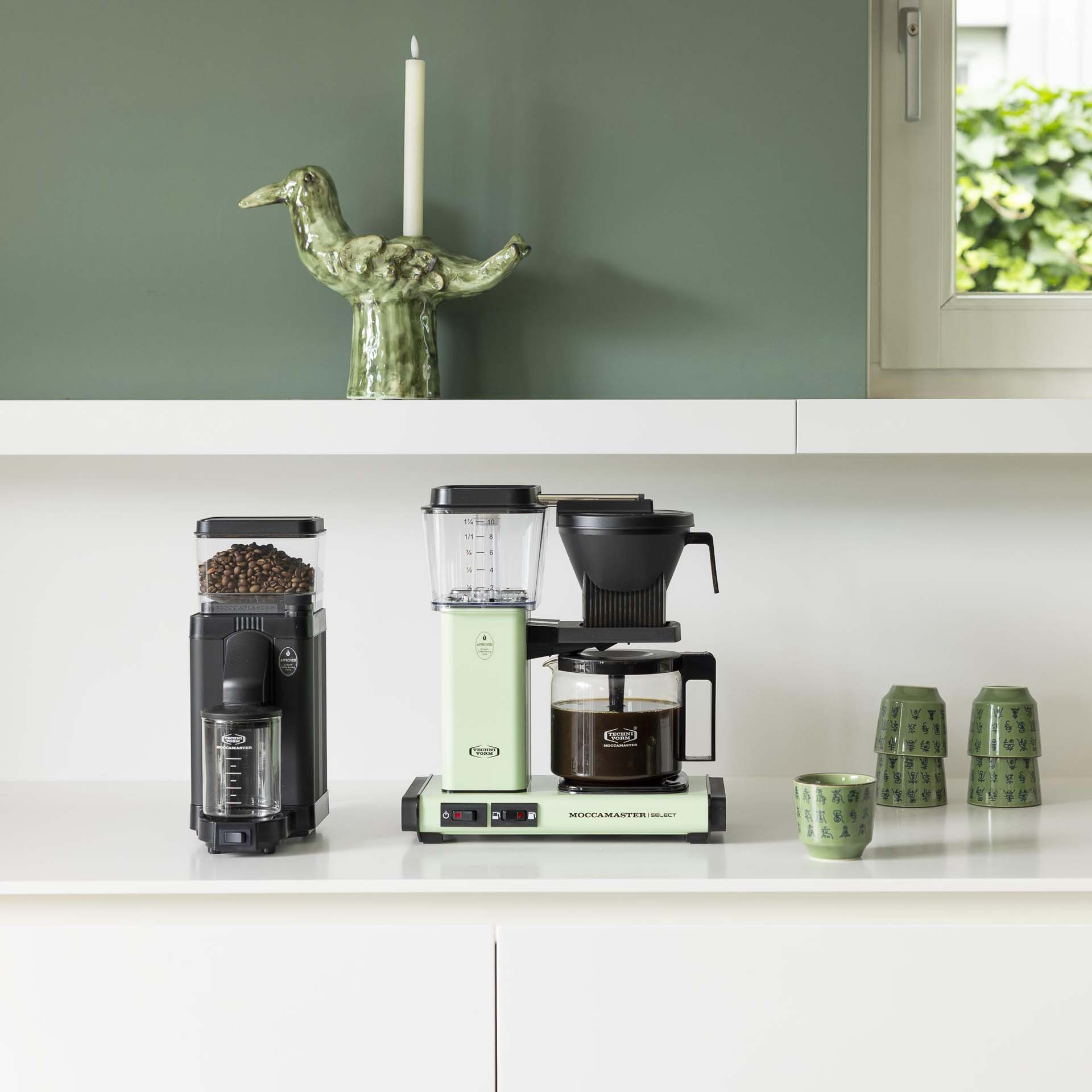 Ekspres do kawy ze zbiornikiem na ziarna i filiżankami na białym blacie.