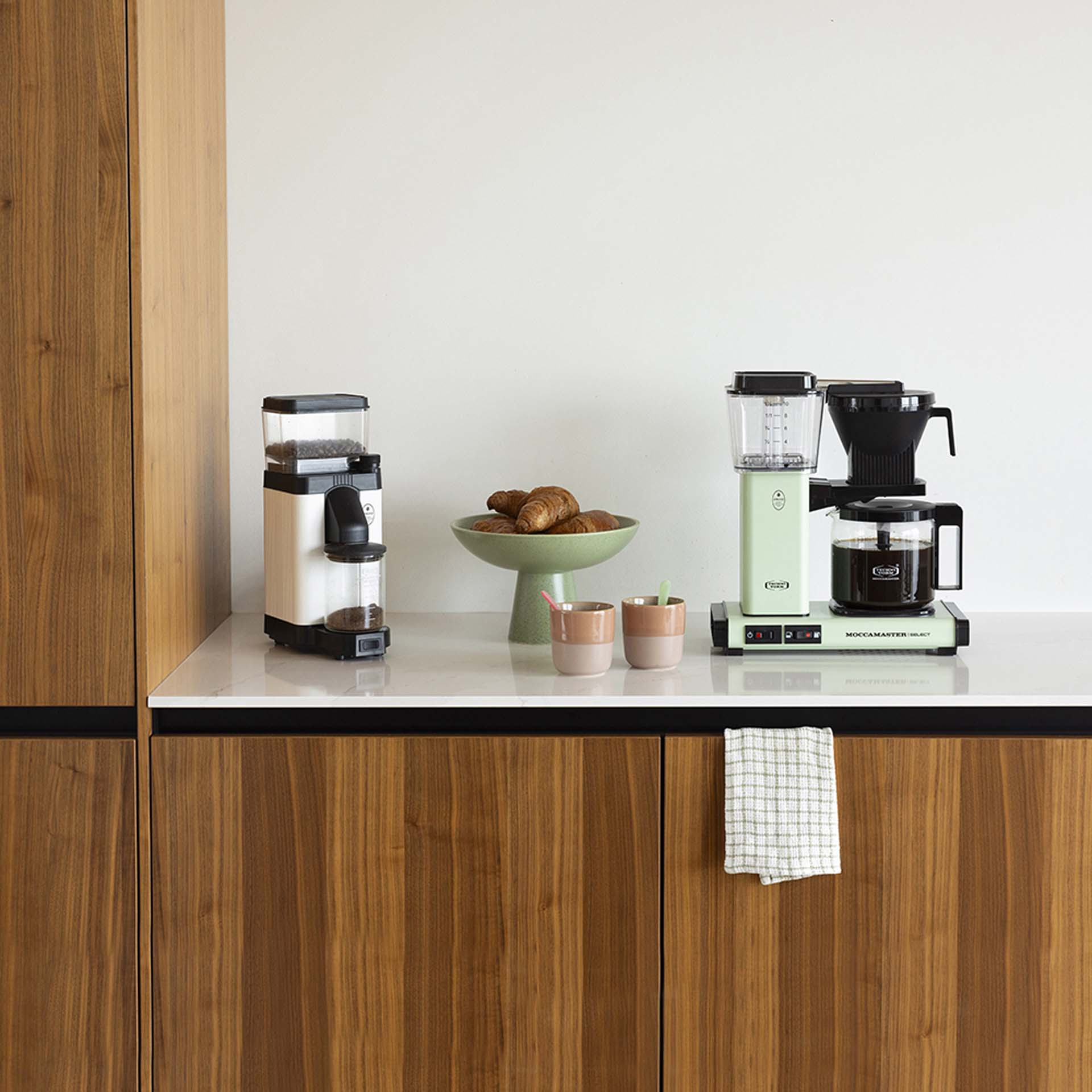 Ekspres do kawy, młynek, rogaliki w misce, dwa kubki na marmurowym blacie.