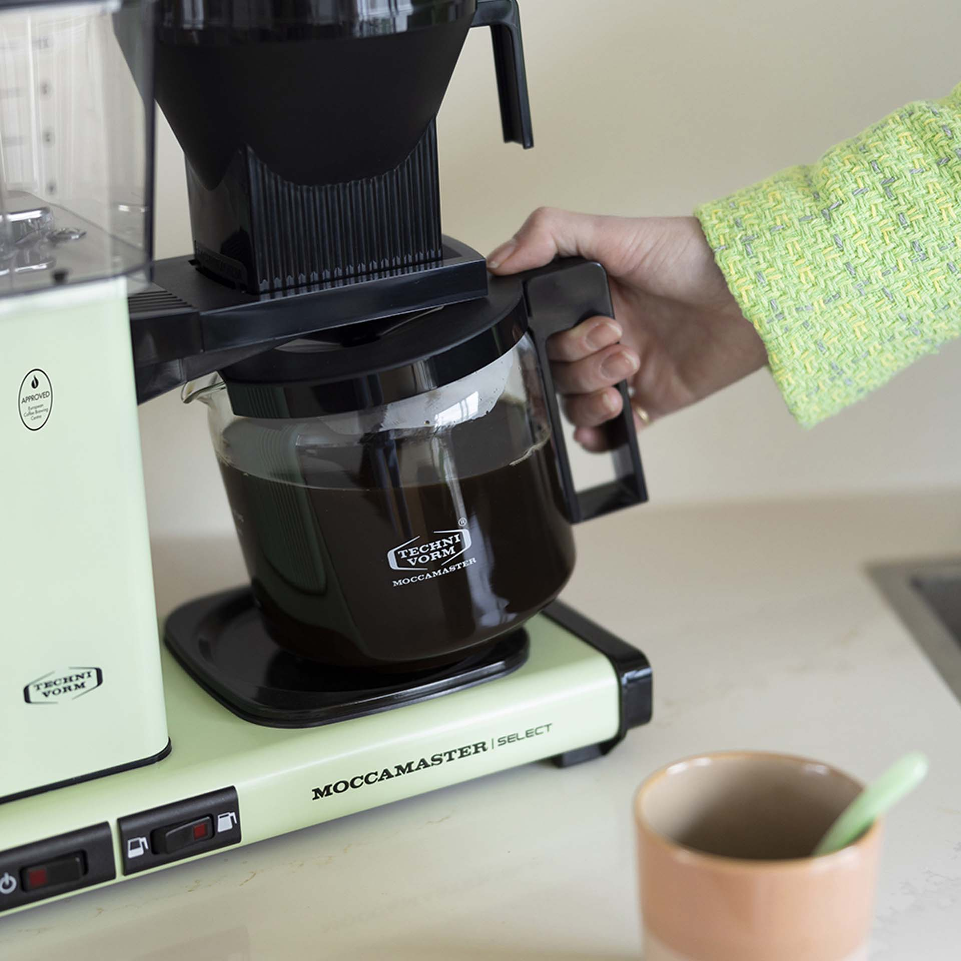 Ręka wyjmuje dzbanek kawy z jasnozielonego ekspresu do kawy.