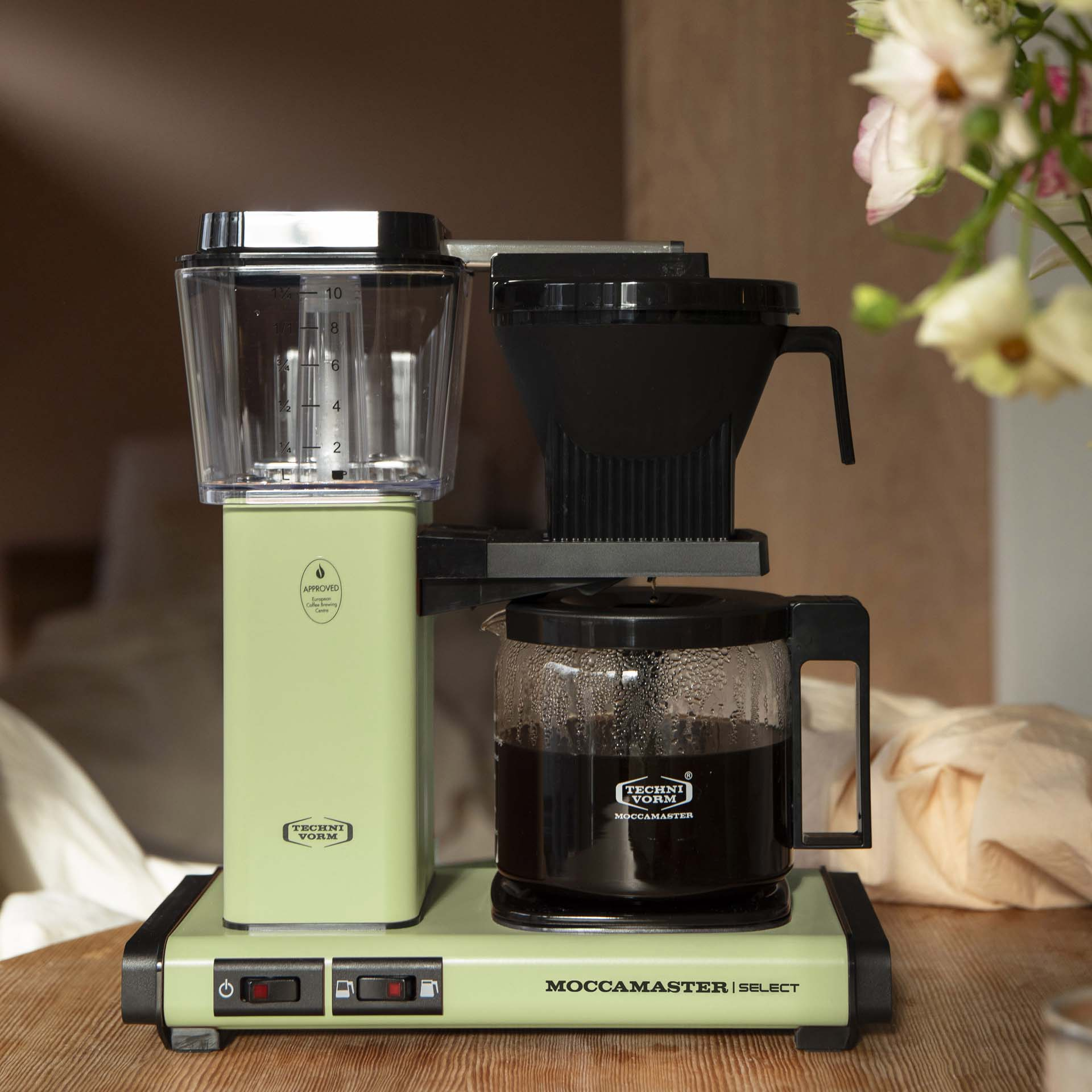 Zielony ekspres do kawy Moccamaster z pełną szklaną karafką stoi na drewnianym stole.