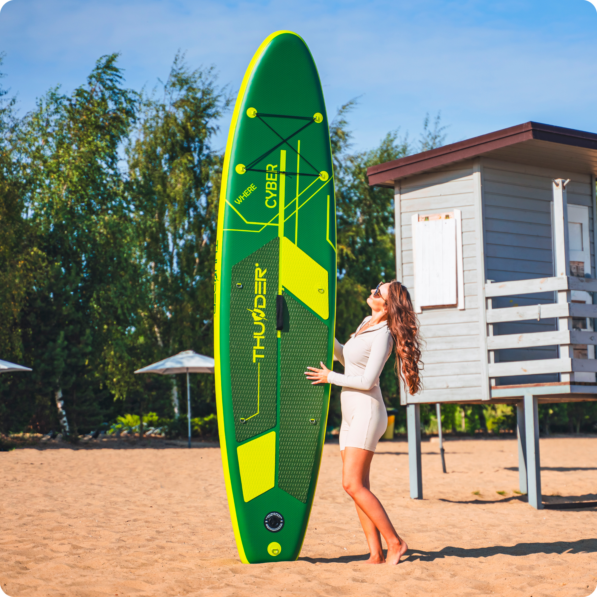 Kobieta na piaszczystej plaży trzyma zielono-żółtą deskę SUP przy wieży ratownika.