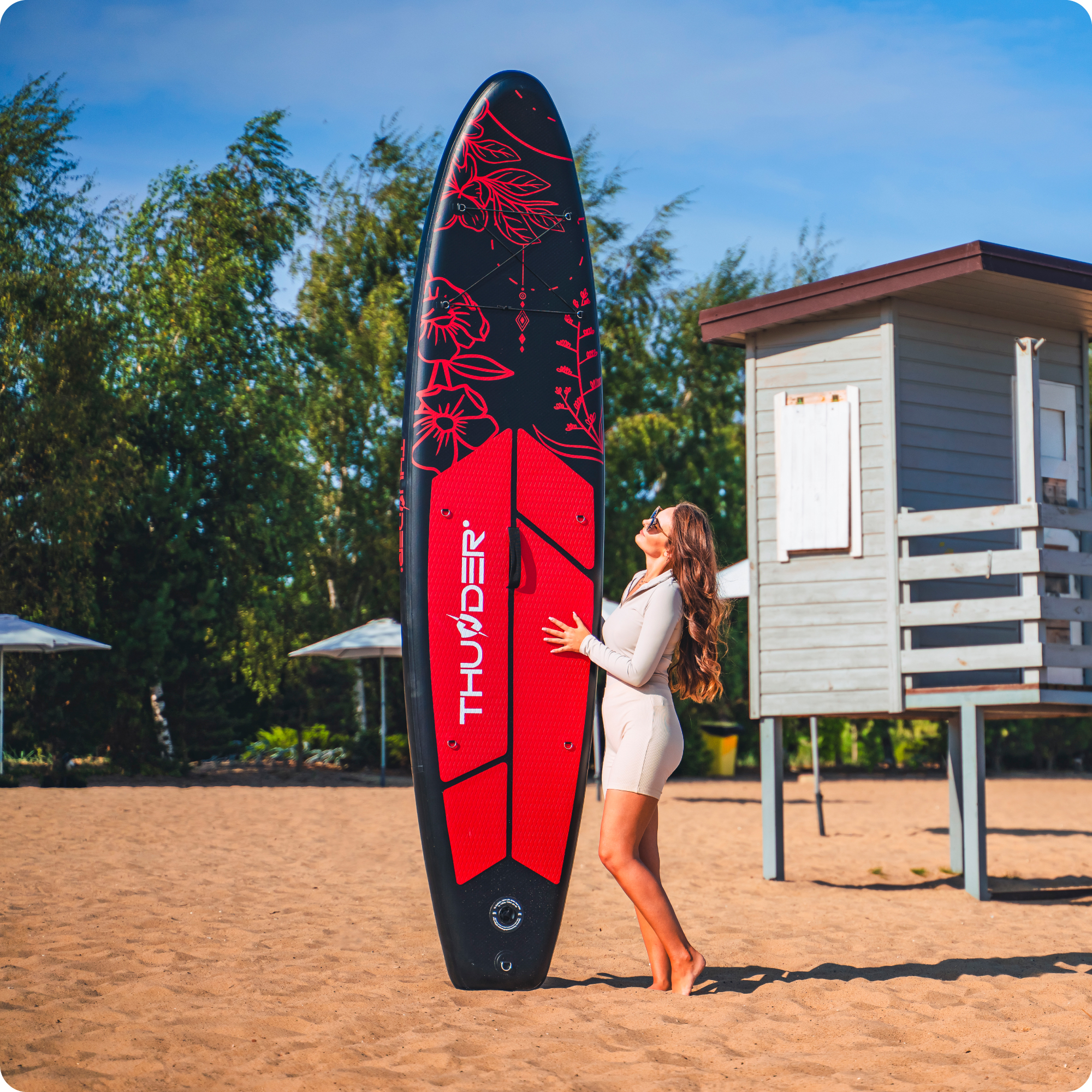 Kobieta stoi na plaży obok czarnej deski SUP z czerwonymi wzorami kwiatowymi.