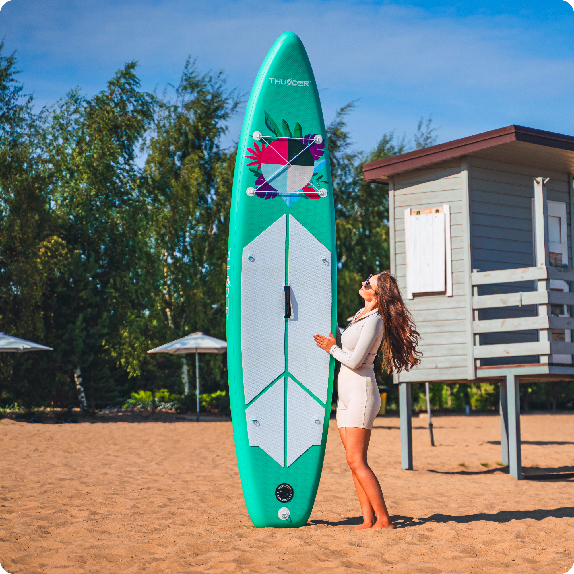 Kobieta stoi na plaży obok turkusowej deski SUP.