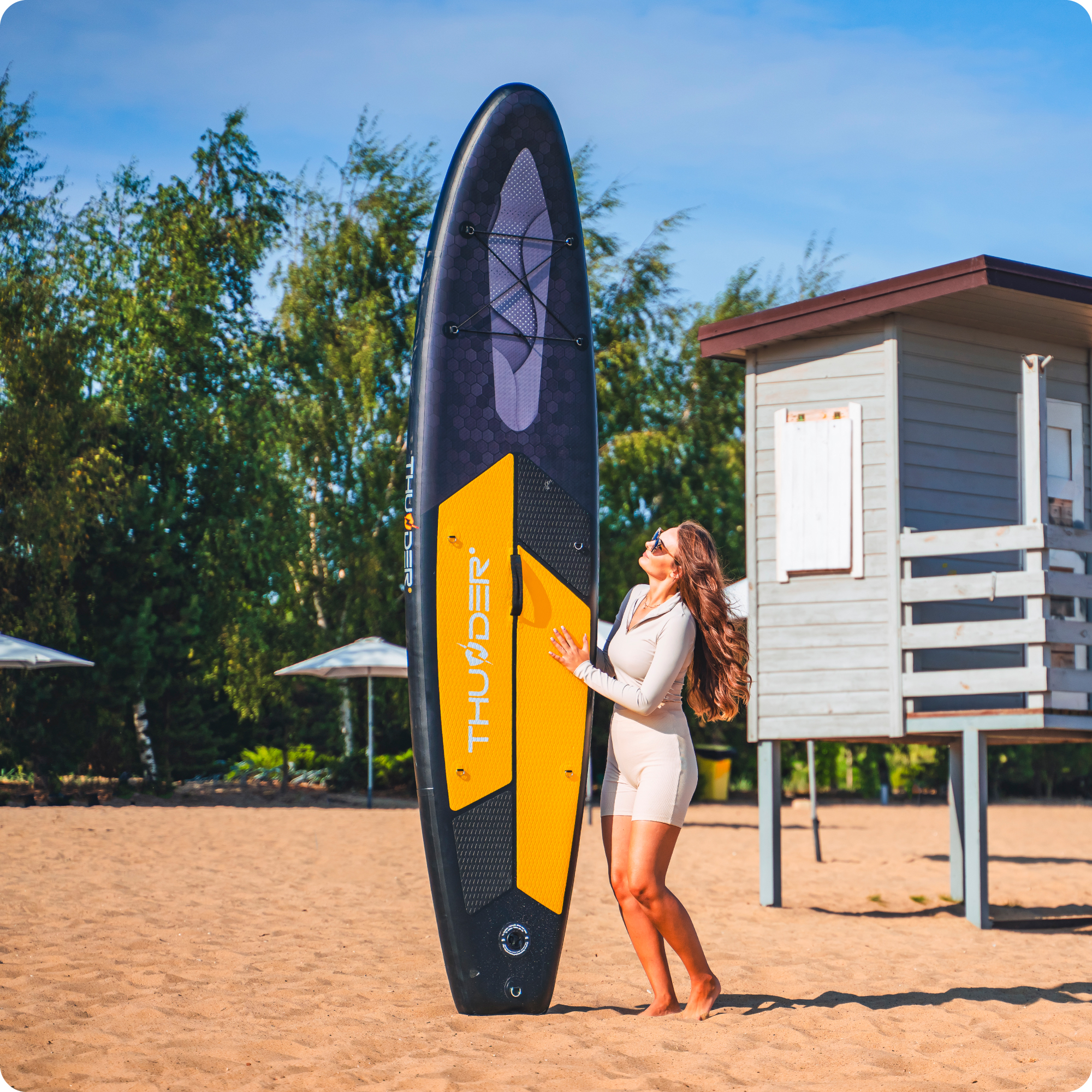 Kobieta z deską SUP na plaży, w tle wieża ratownicza.