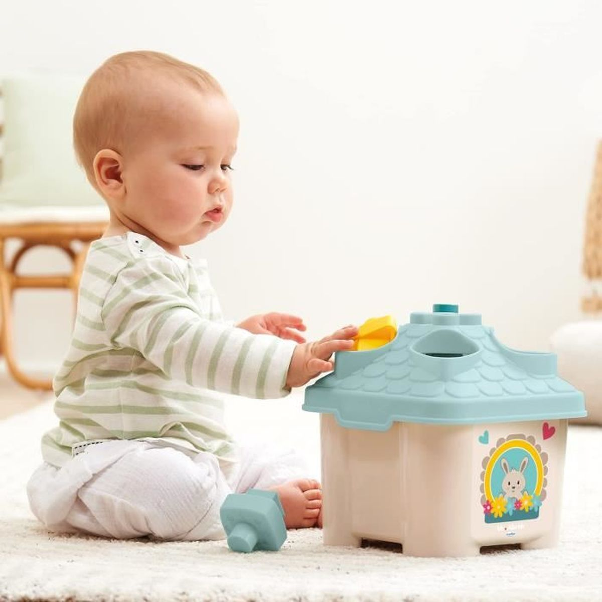 Ein Baby im gestreiften Hemd spielt mit einem Formsortierhaus-Spielzeug auf einem Teppich.