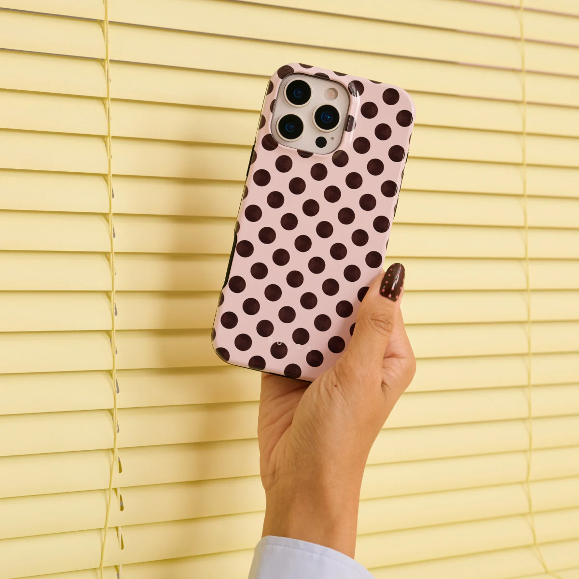 Een hand houdt een roze telefoonhoesje met bruine stippen voor gele jaloezieën.