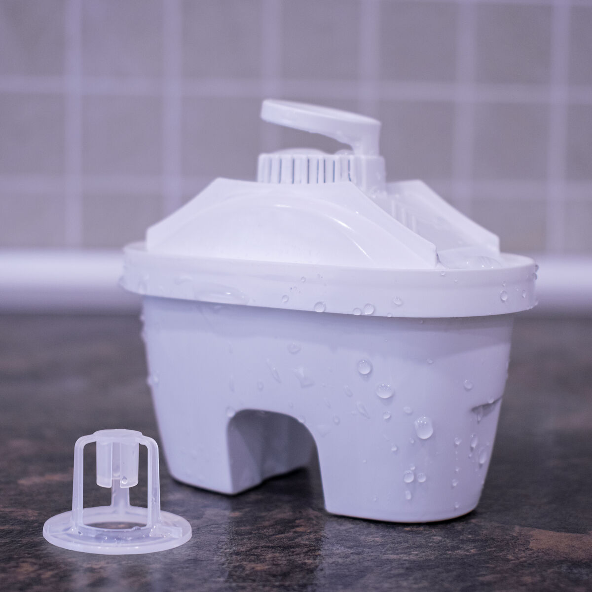 Ein weißer Wasserfilter mit Wassertropfen darauf. Ein kleiner Plastikteil liegt daneben.