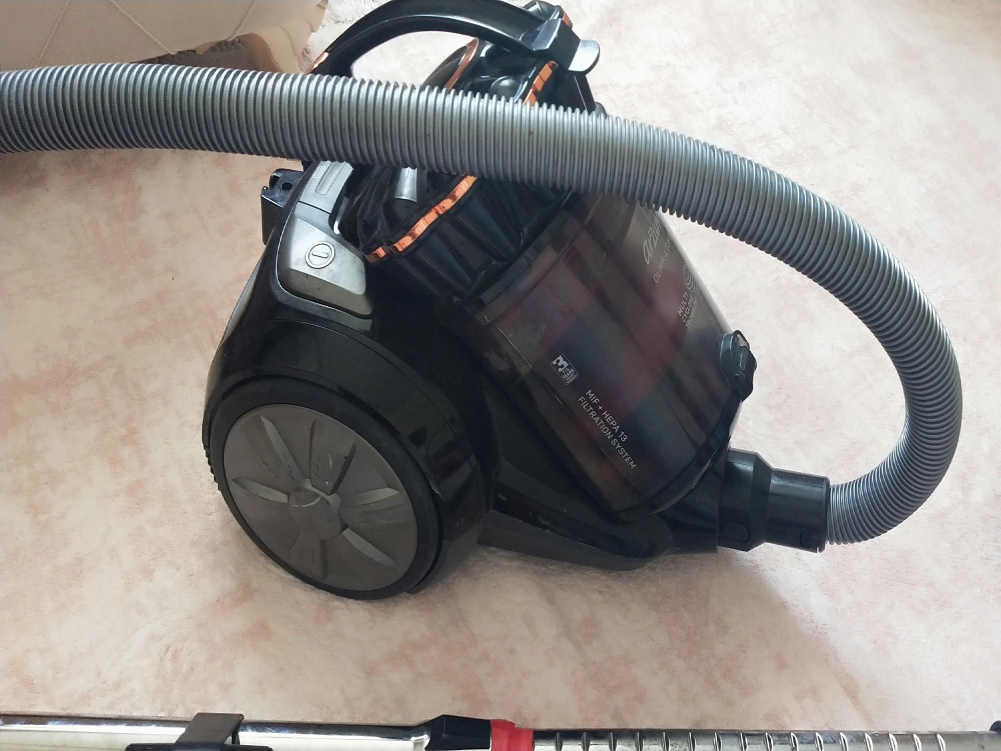 Hortumlu ve tutamaklı siyah bir elektrikli süpürge açık renk halının üzerinde duruyor.