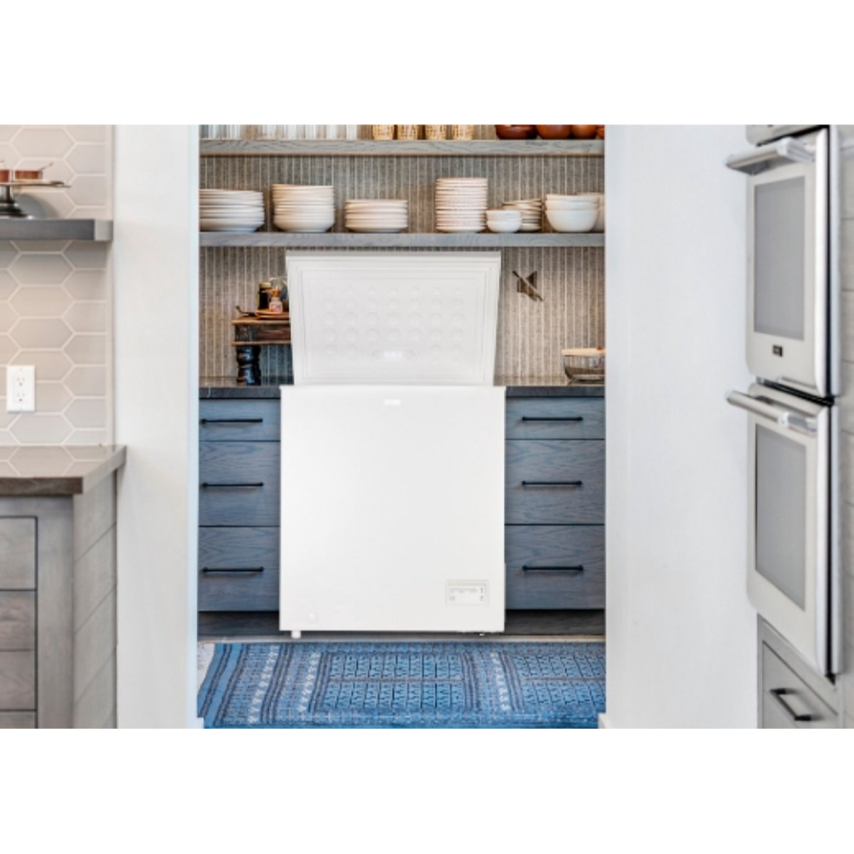 Biała zamrażarka skrzyniowa z otwartą pokrywą stoi na niebieskim dywanie.