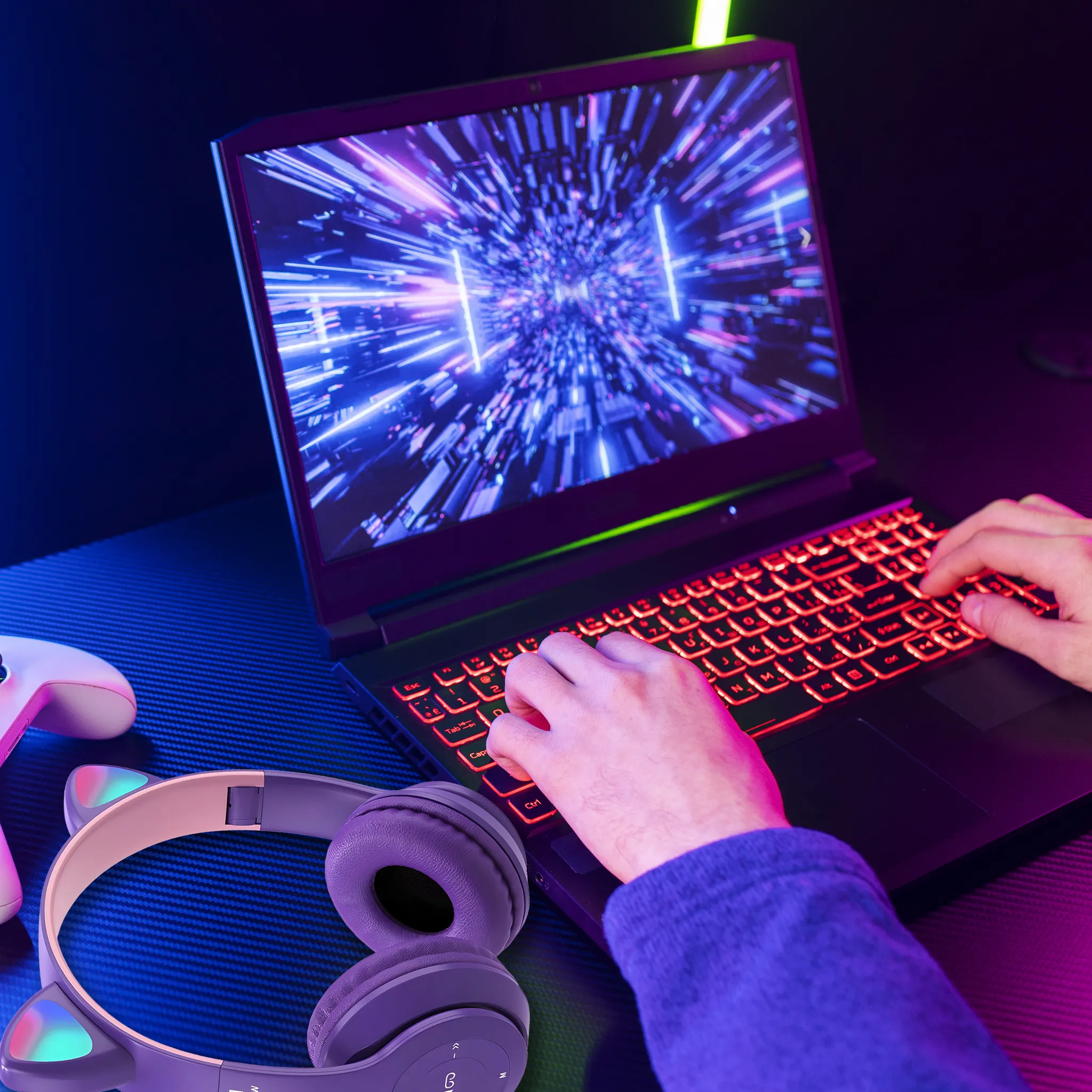 Person tippt auf Tastatur eines Laptops mit beleuchteter Tastatur. Kopfhörer und Controller liegen daneben.