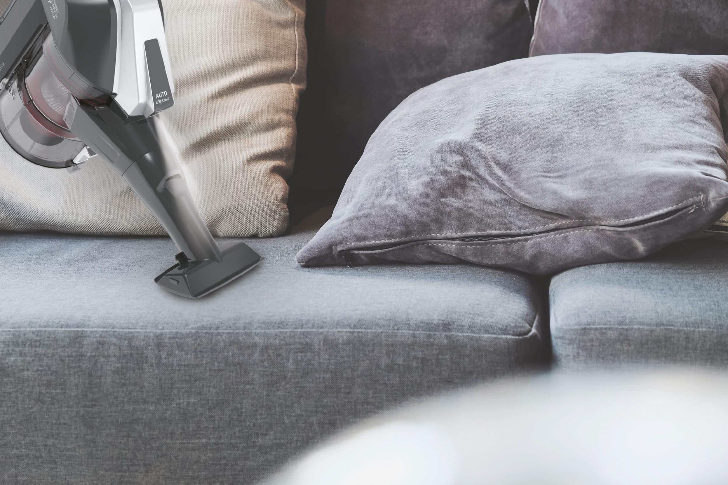 Ein Handstaubsauger reinigt ein graues Sofa mit Kissen.