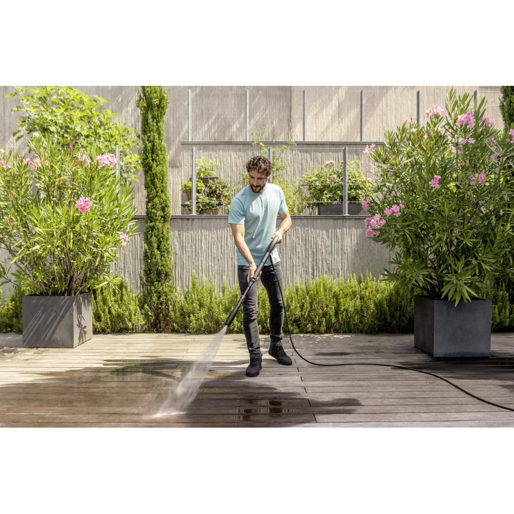 Mężczyzna czyści drewniany taras myjką ciśnieniową obok kwitnących roślin.