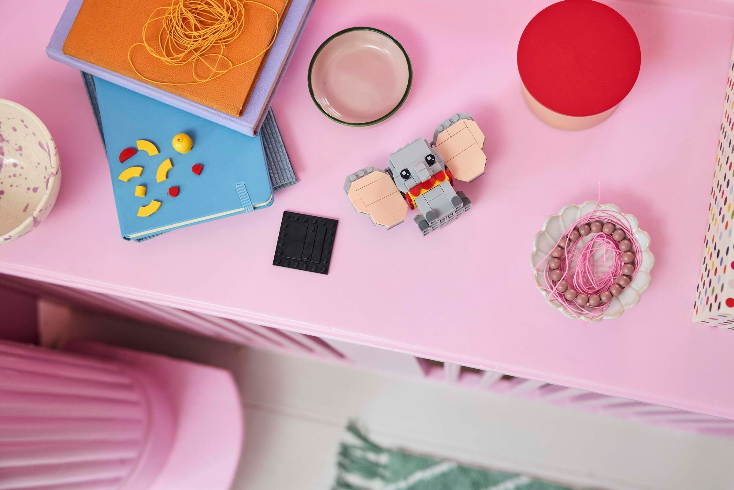 Grauer Lego-Elefant mit rosa Ohren auf einem rosa Tisch mit Notizbüchern und Perlen.