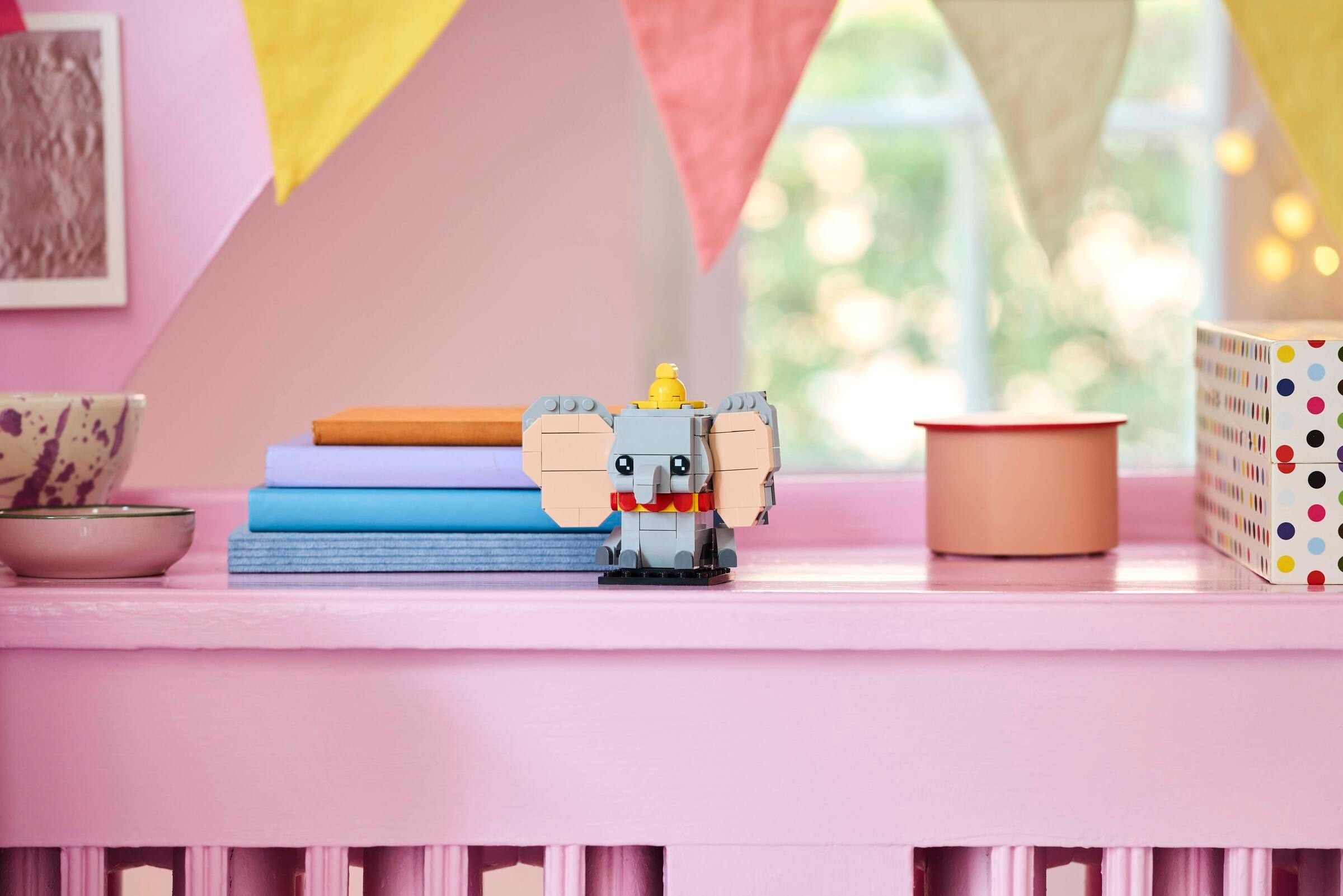 Grauer Lego-Elefant mit gelbem Hut steht auf einem rosafarbenen Regal neben Büchern und Behältern.