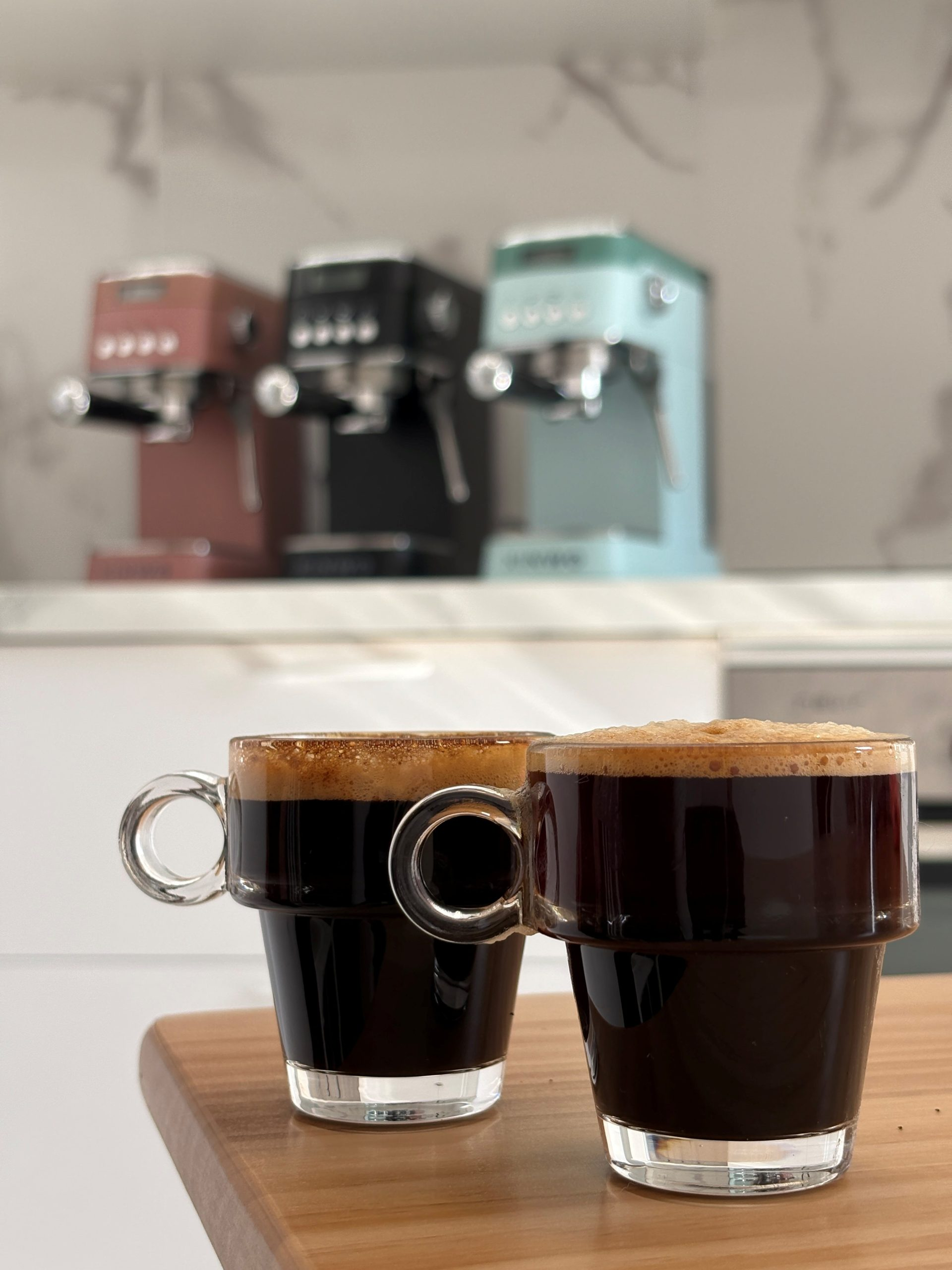 Dos vasos de espresso con crema sobre mesa de madera. Tres máquinas de café al fondo.