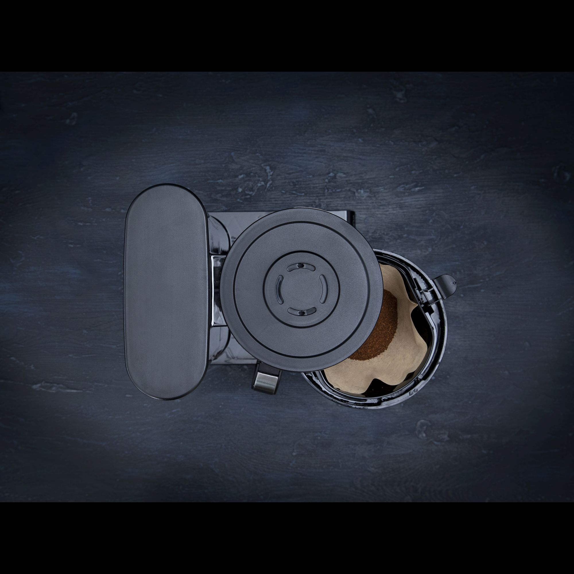 A black coffee maker with filter and coffee grounds on a dark background.