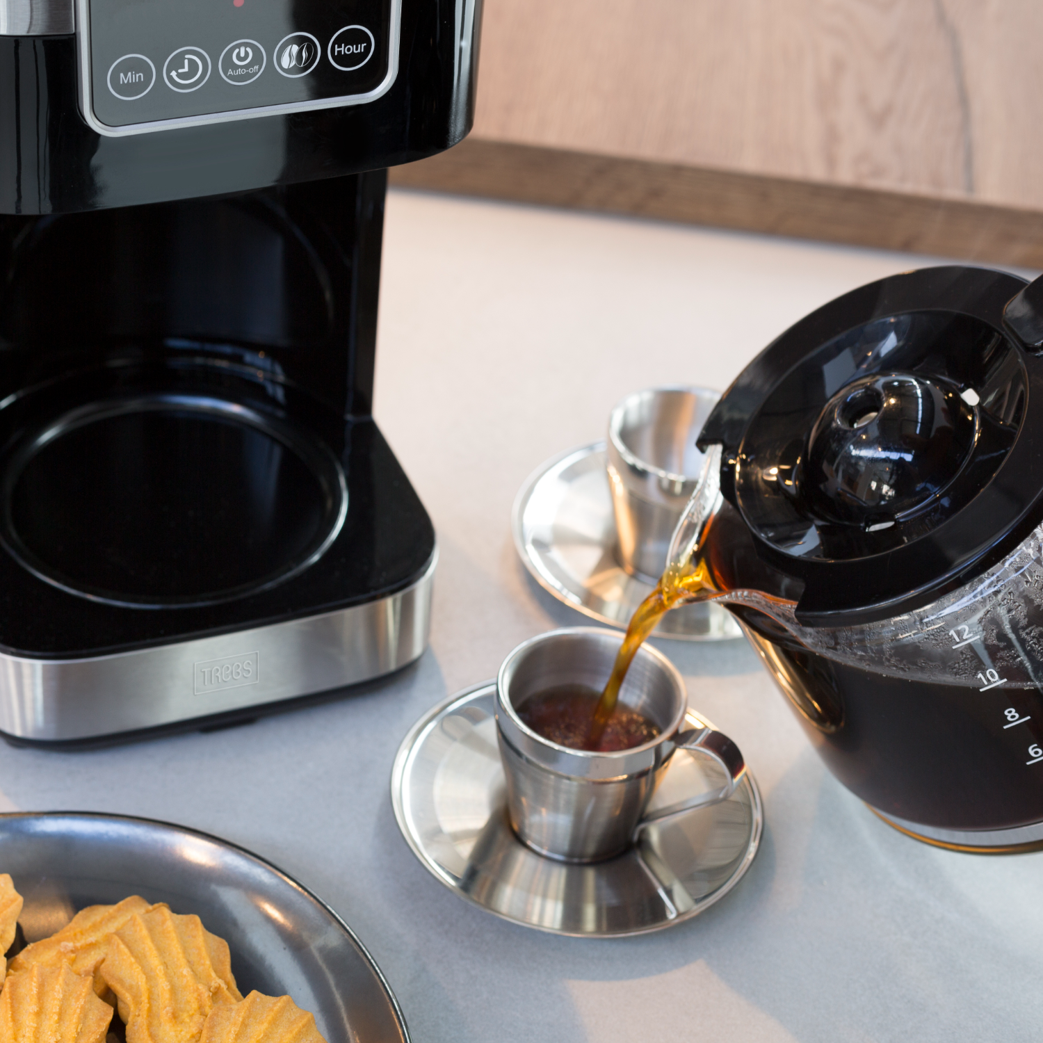 Kaffeekanne gießt Kaffee in eine Tasse, neben Keksen und einer Kaffeemaschine.