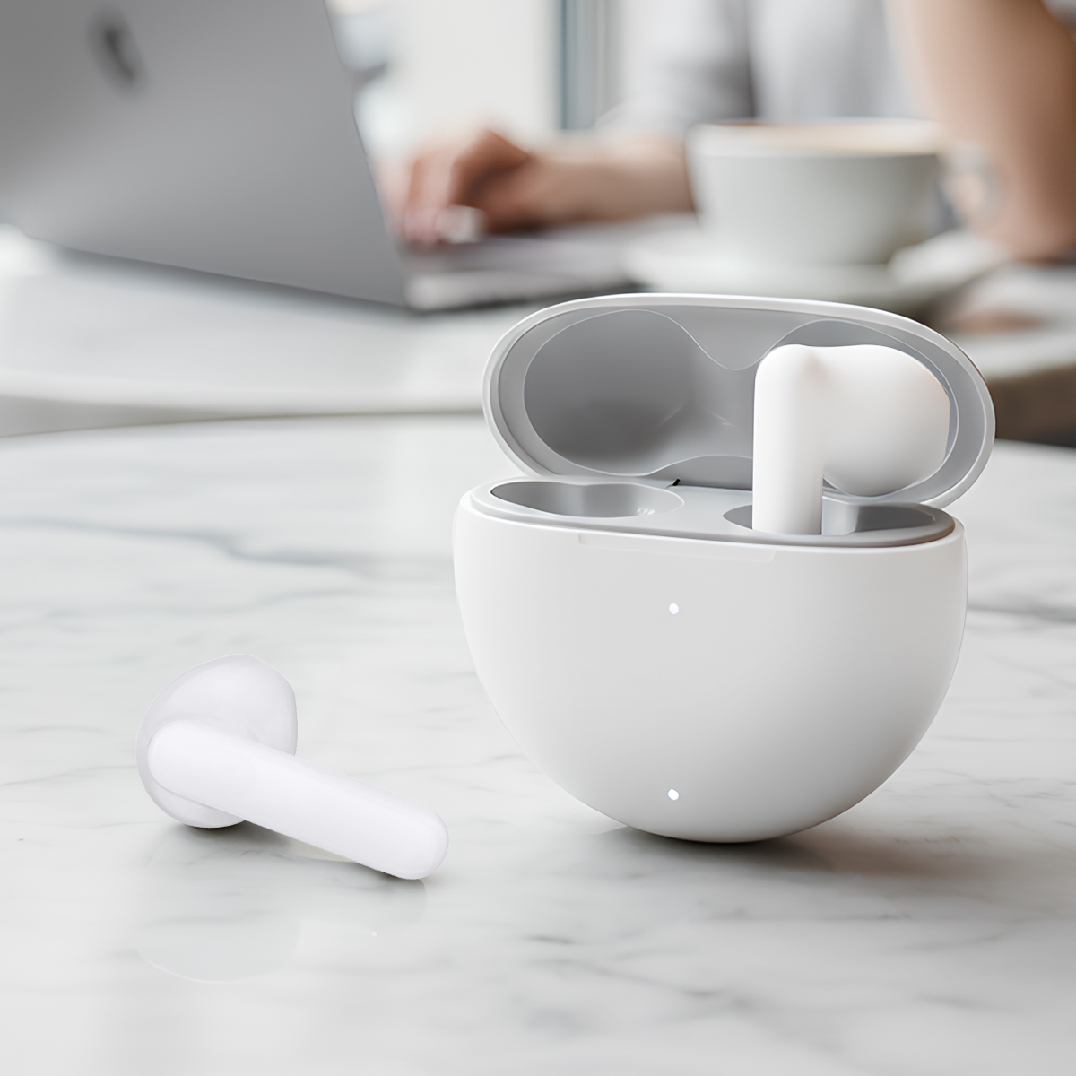 Białe słuchawki i etui ładujące na marmurowym stole, laptop i kawa w tle.