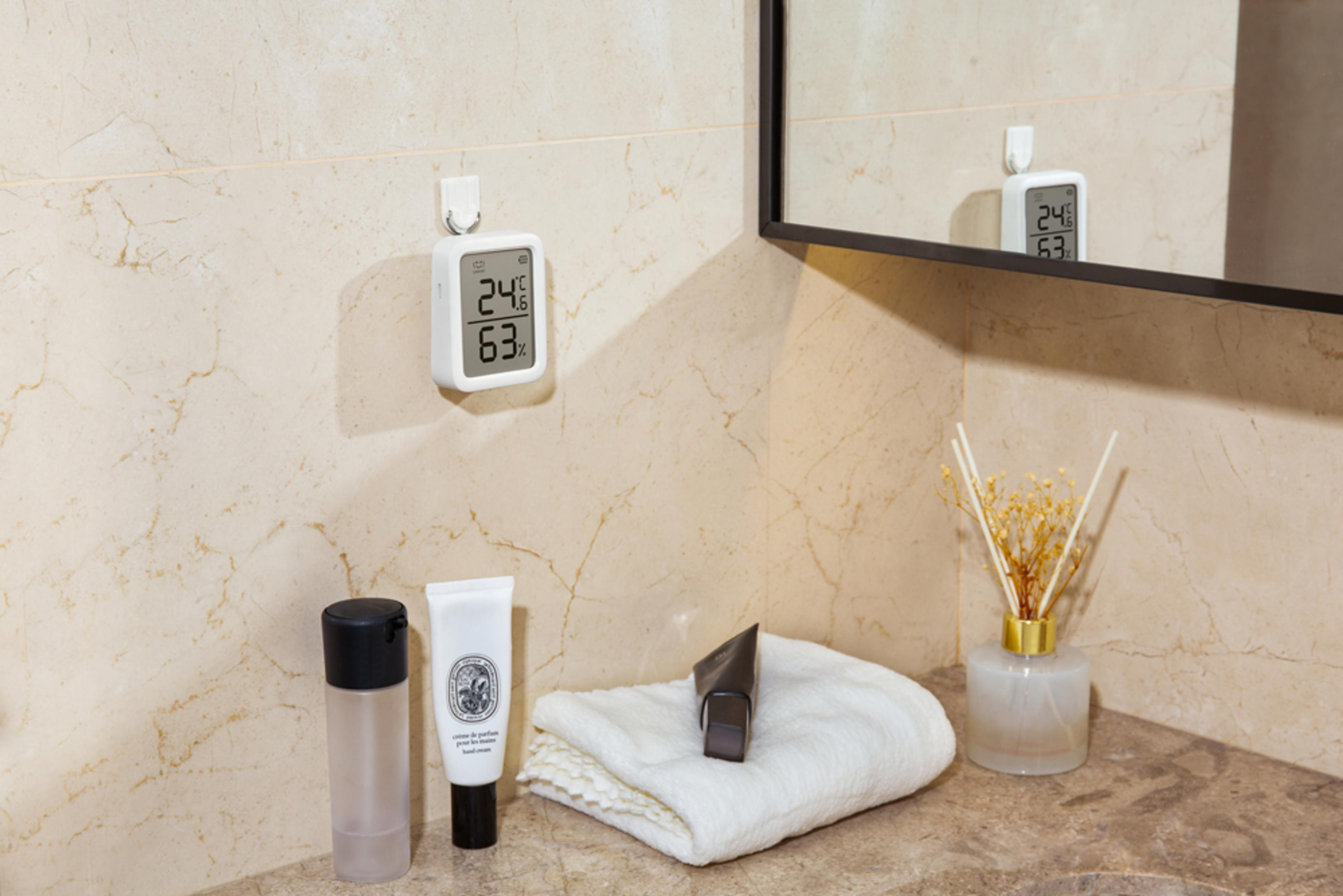 Ein digitaler Thermometer/Hygrometer hängt an einer gefliesten Wand neben Toilettenartikeln.