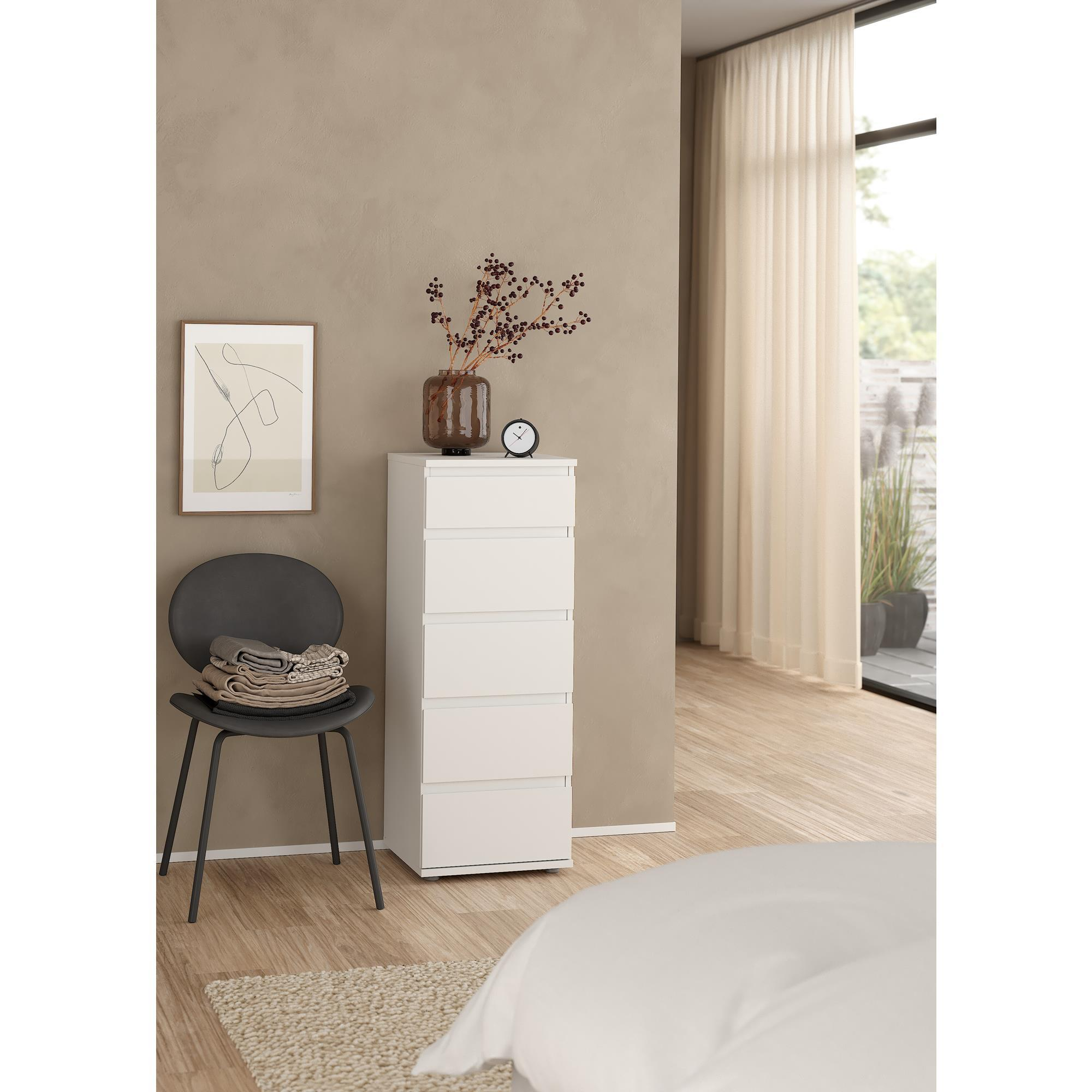 Un dormitorio minimalista con cómoda blanca, silla oscura con ropa y ventana.