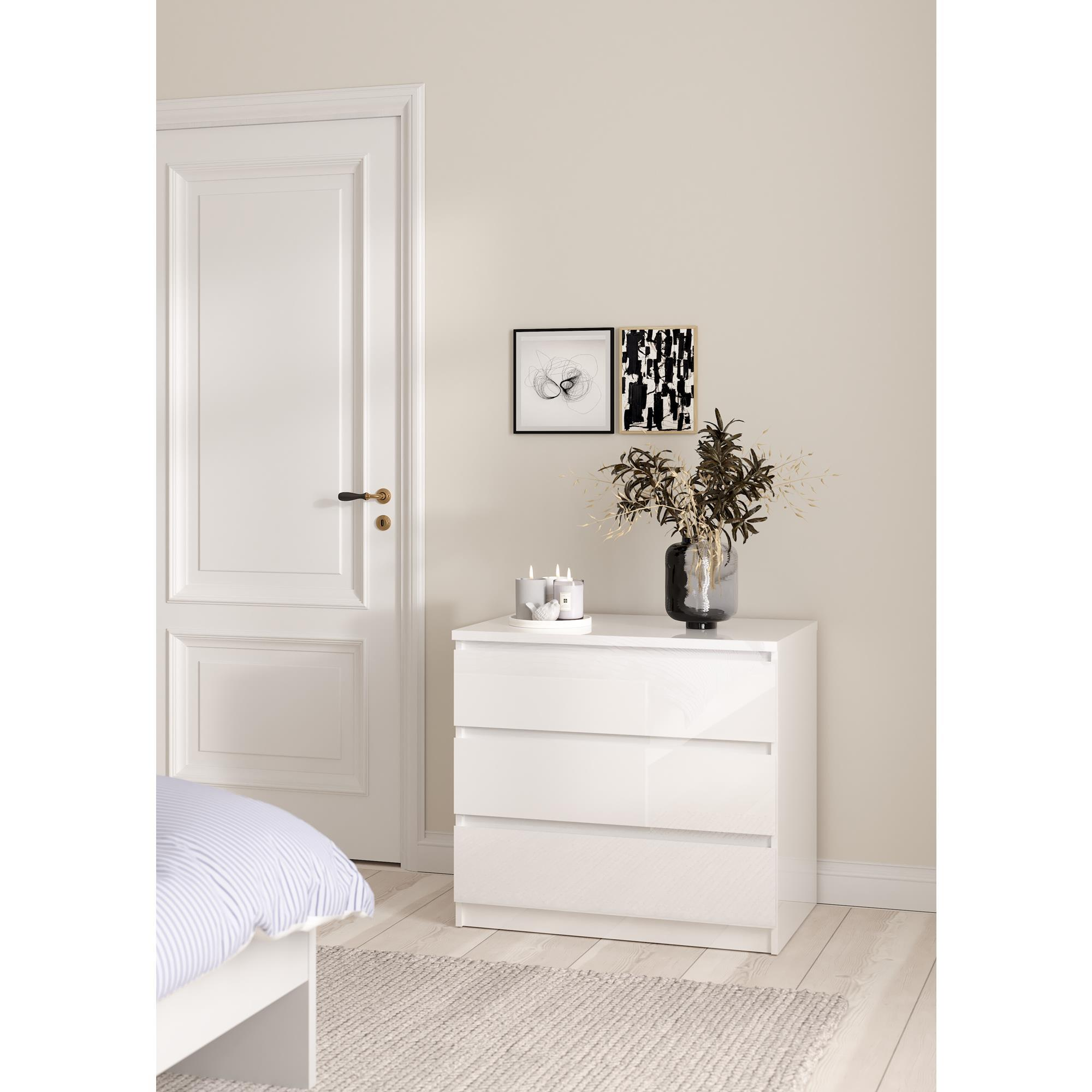 Cómoda blanca con cajones junto a una cama, frente a una puerta blanca.