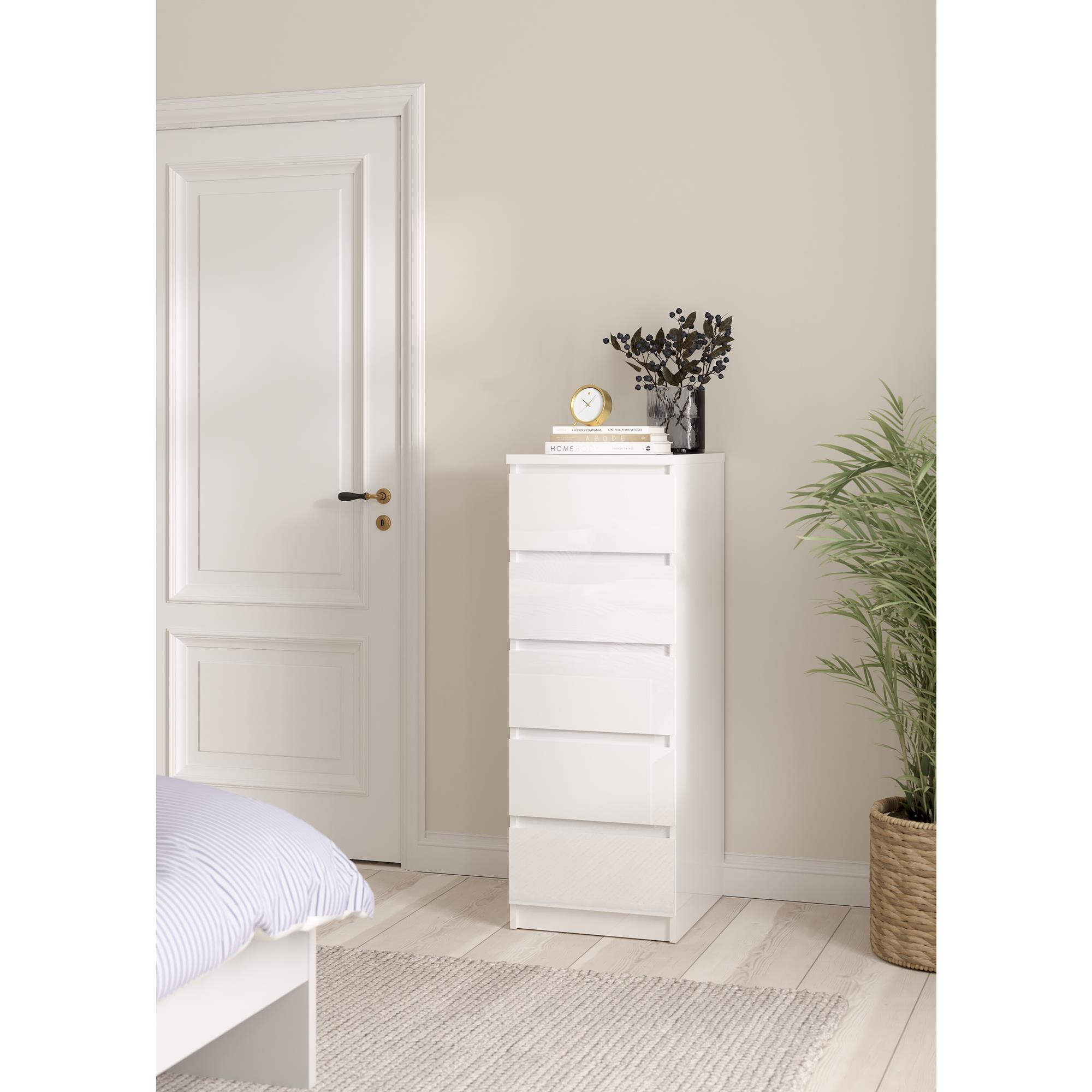 Una cómoda blanca está al lado de una cama. Una puerta y una planta están al fondo.