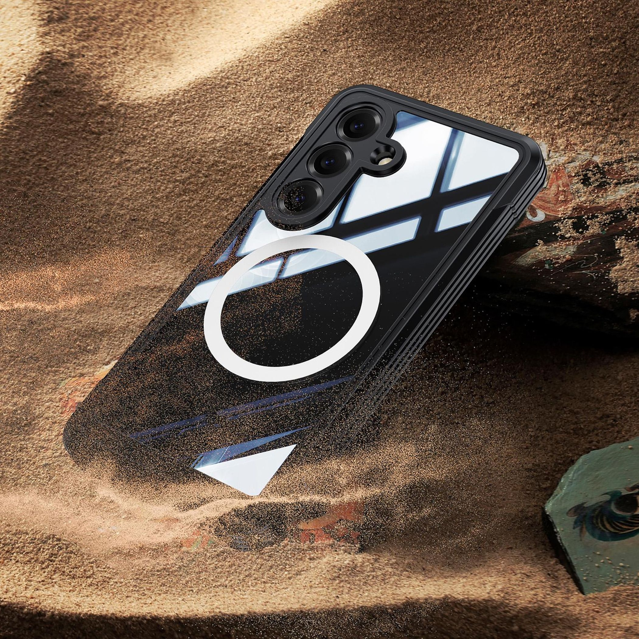 Un teléfono negro con anillo blanco descansa sobre arena. Módulo de cámara visible.