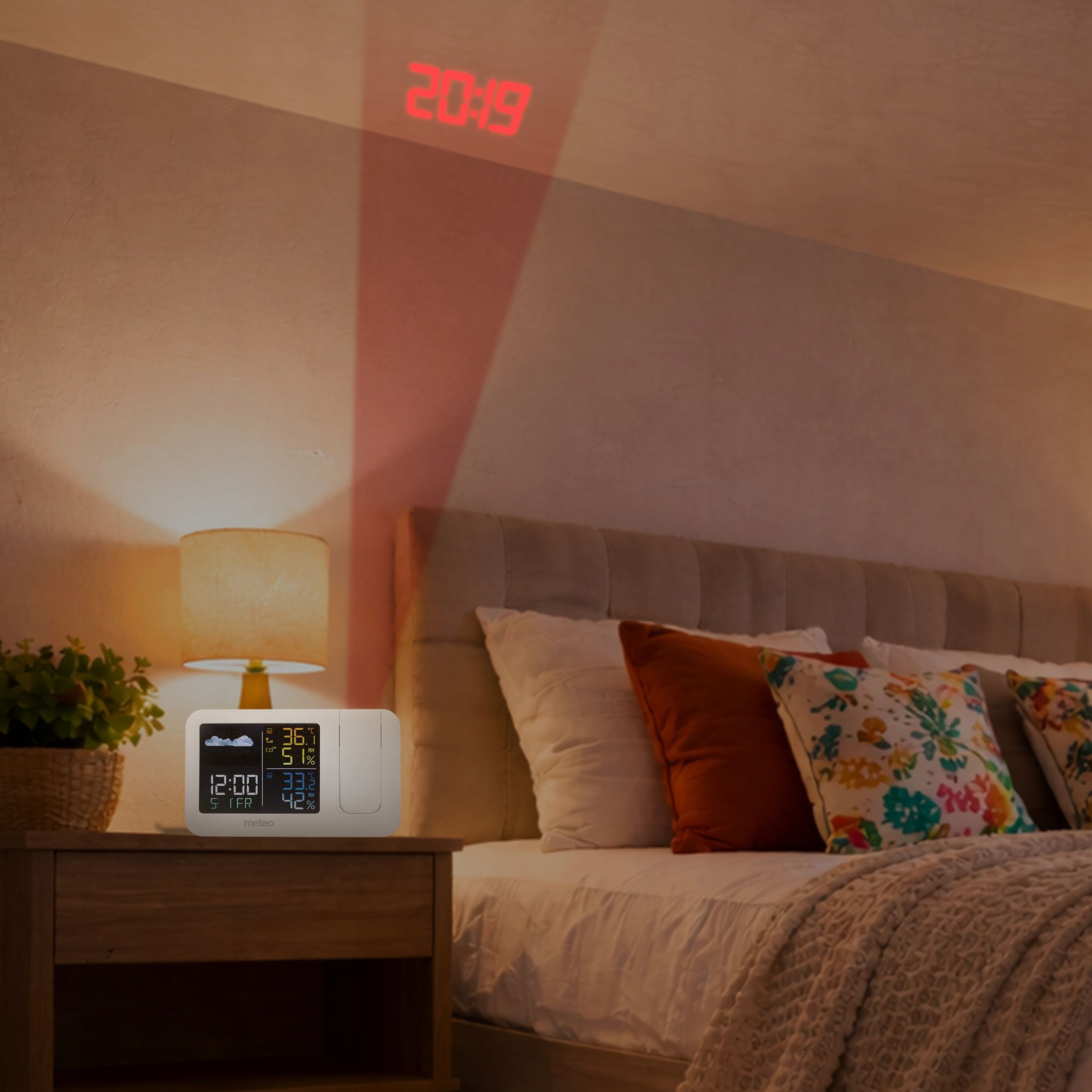 Stolik nocny ze stacją pogodową i lampą obok łóżka z kocem.