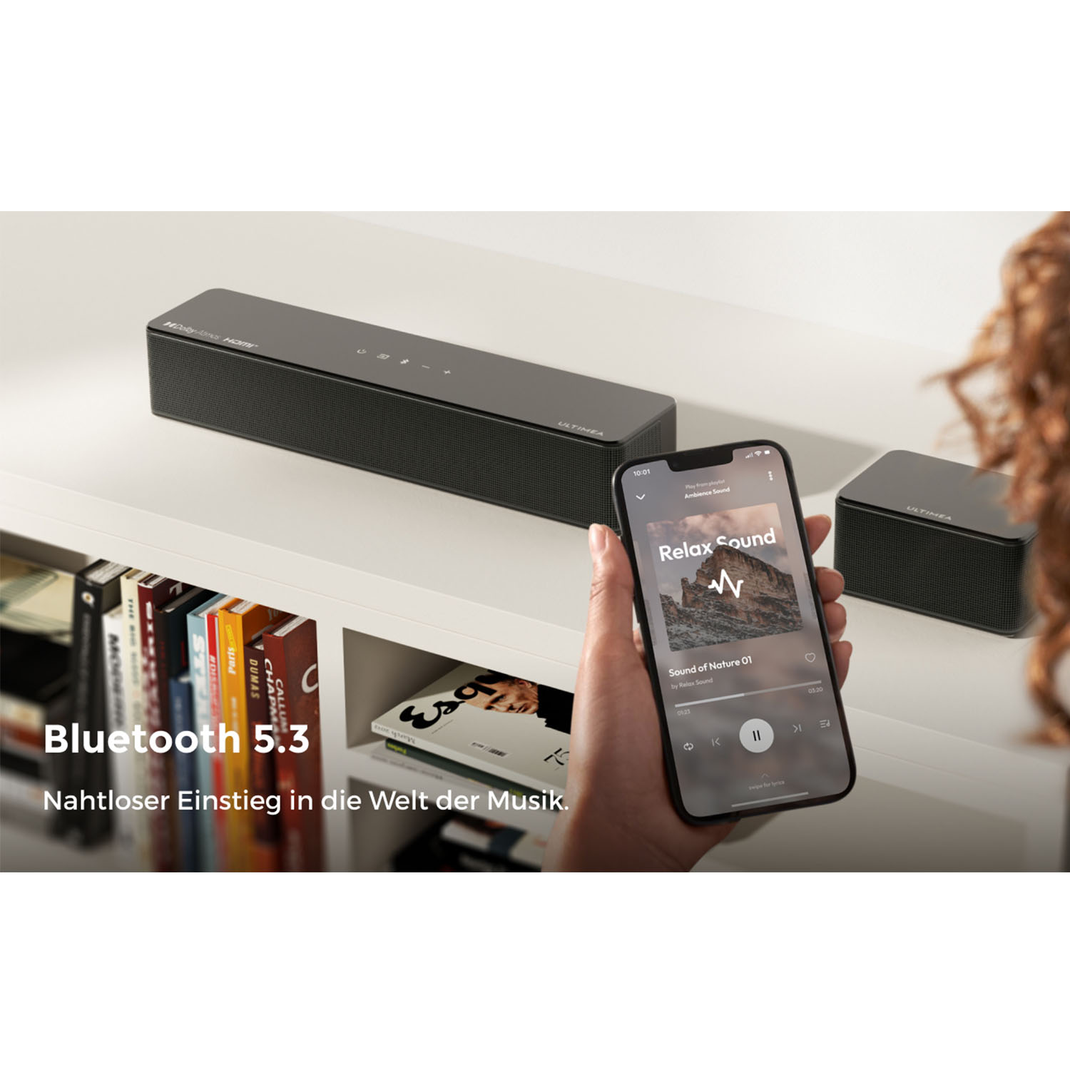 Eine Person hält ein Smartphone mit Musik-App, zwei schwarze Lautsprecher stehen daneben.