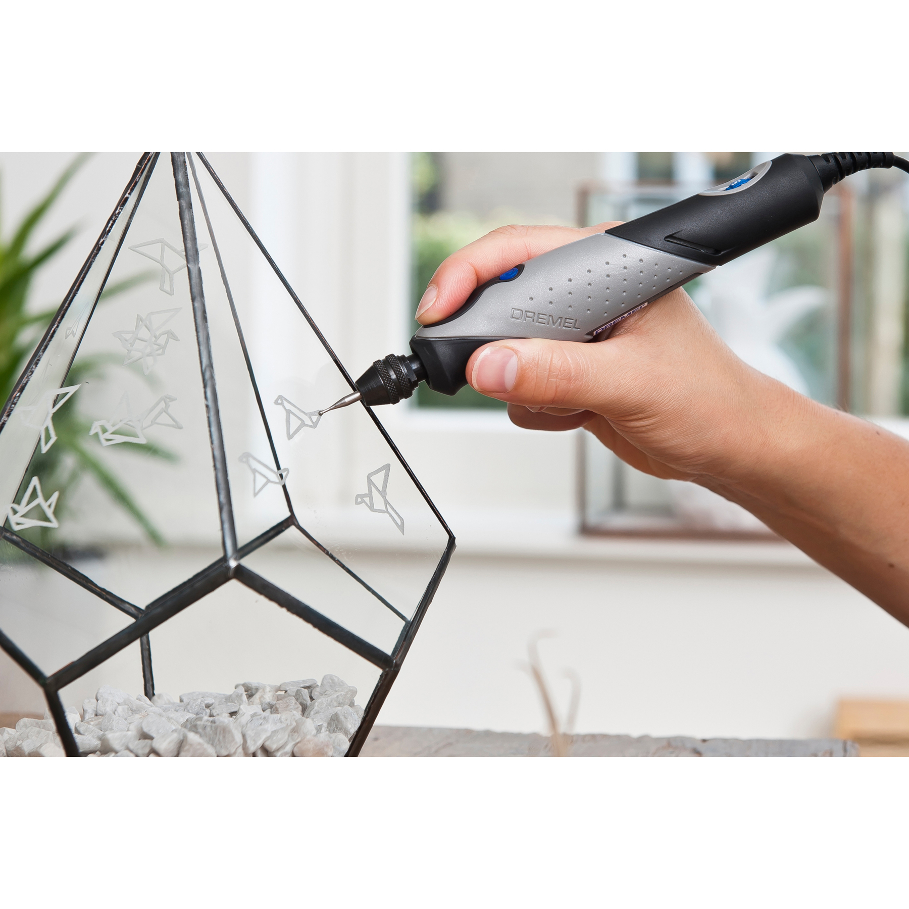 Eine Hand benutzt ein Gravierwerkzeug, um Origami-Vögel auf ein Glasobjekt zu ätzen.