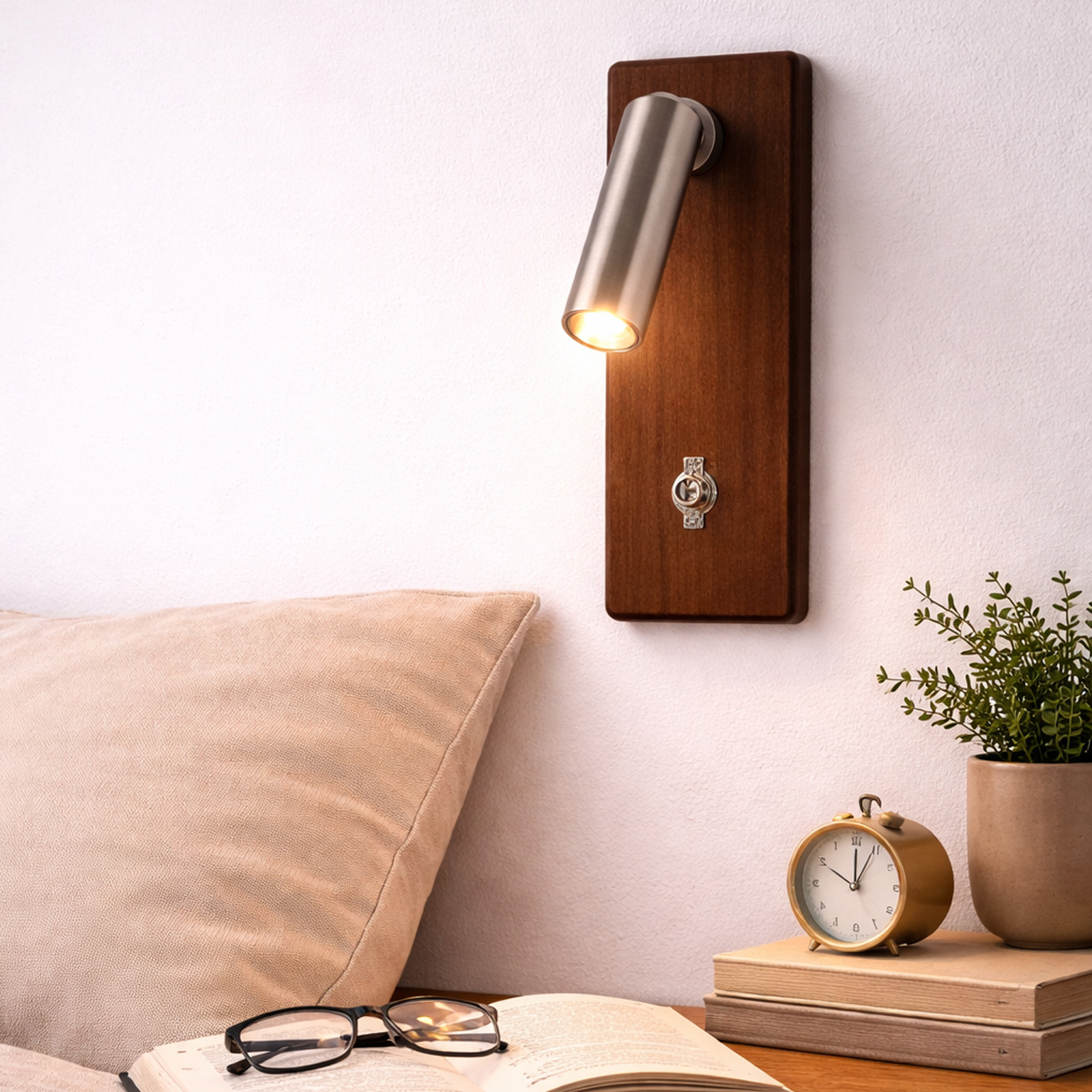 Una mesita de noche con libro, gafas, despertador y planta junto a una cama.