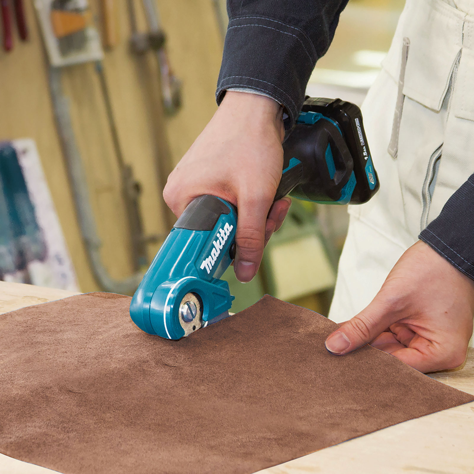 Person schneidet Leder mit blauem Werkzeug. Werkstatt im Hintergrund.