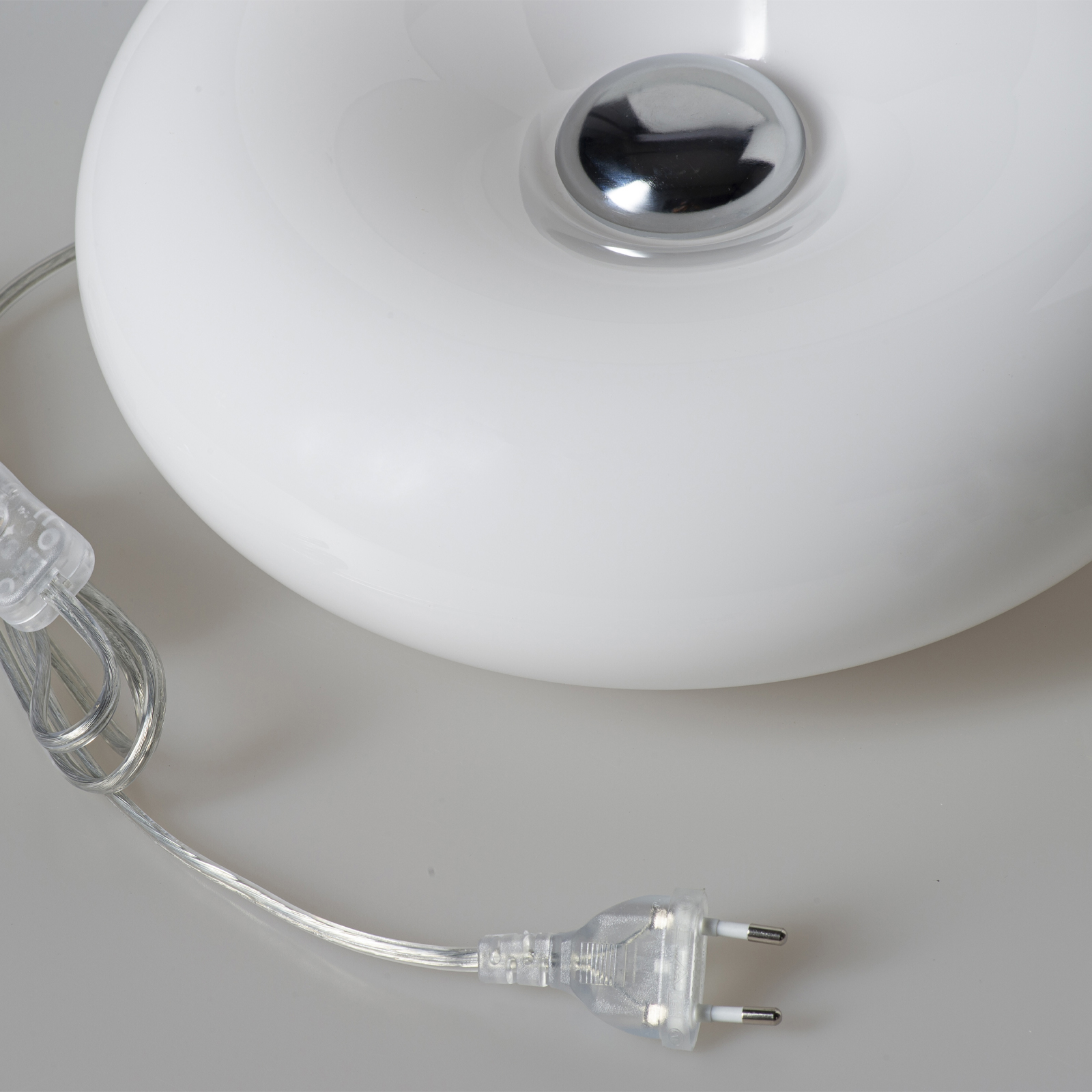 Un objeto blanco, liso y redondo con un botón plateado brillante en la parte superior, y un cable eléctrico transparente con enchufe.