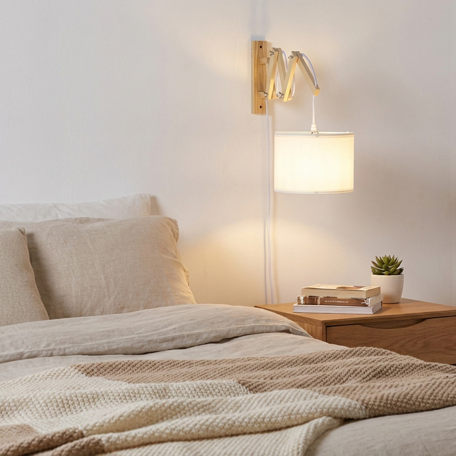 Una mesita de noche con libros y planta junto a una cama con lámpara de pared.