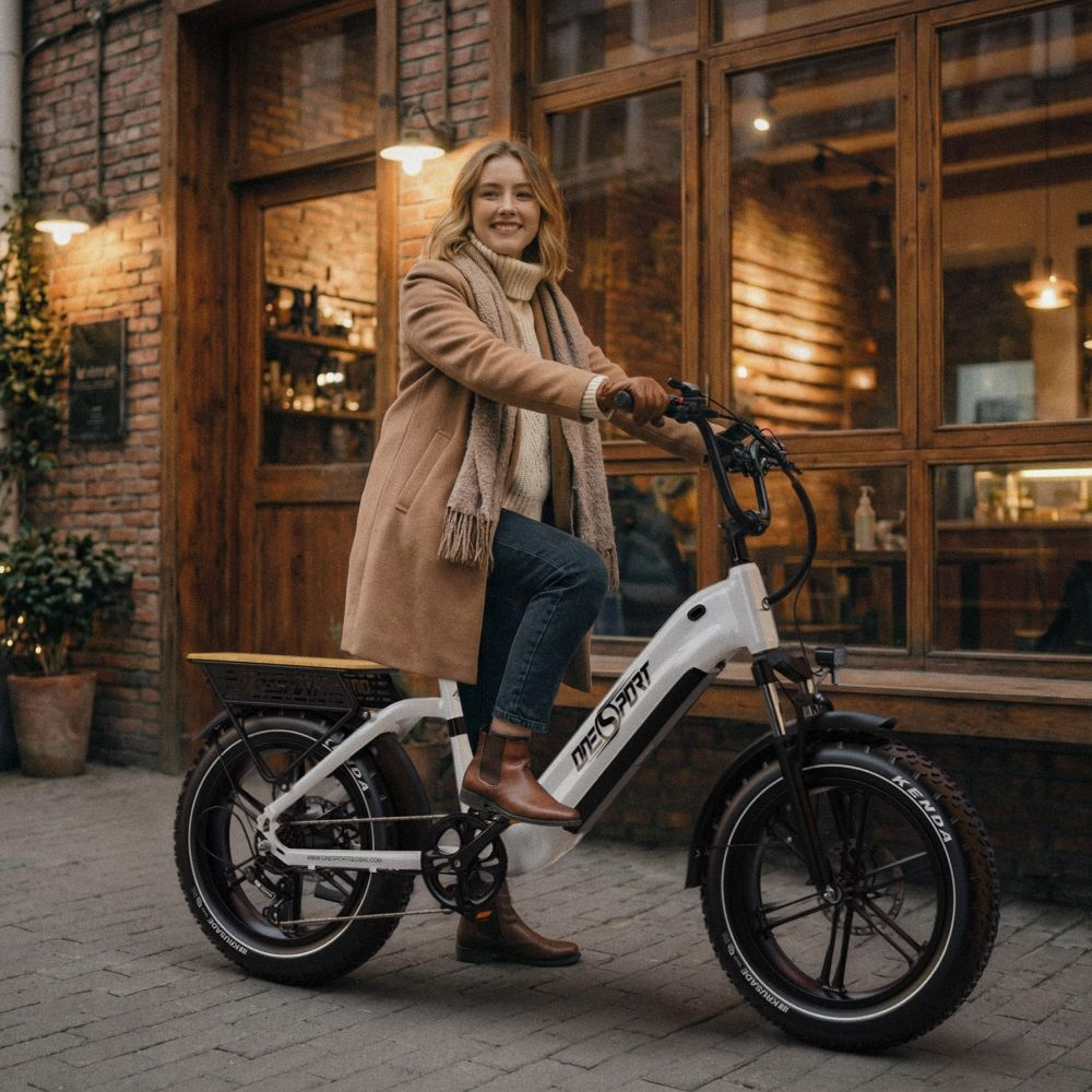 Una donna con cappotto beige guida una e-bike bianca davanti a un edificio di mattoni.