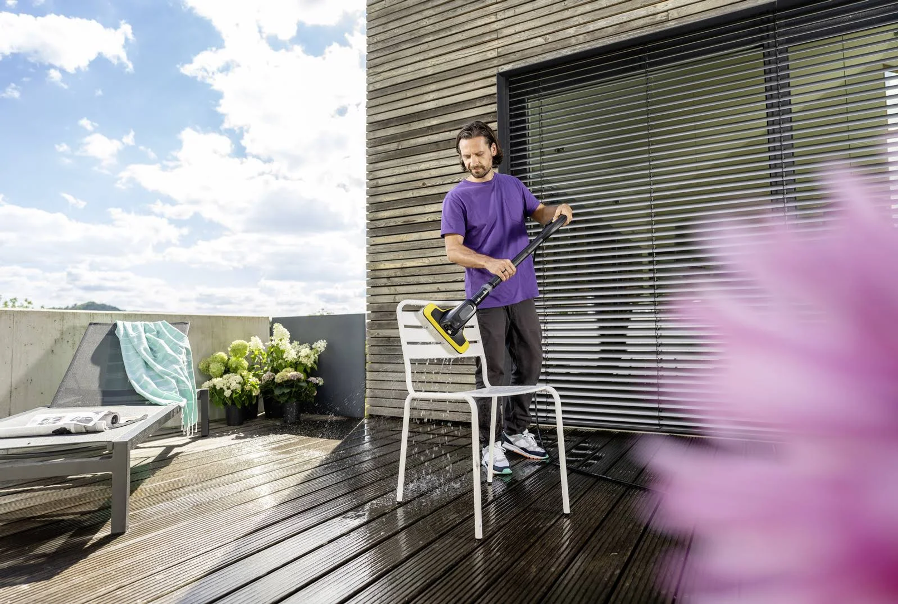 Mann reinigt weiße Stuhlbeine mit einem Hochdruckreiniger auf einer Holzterrasse.