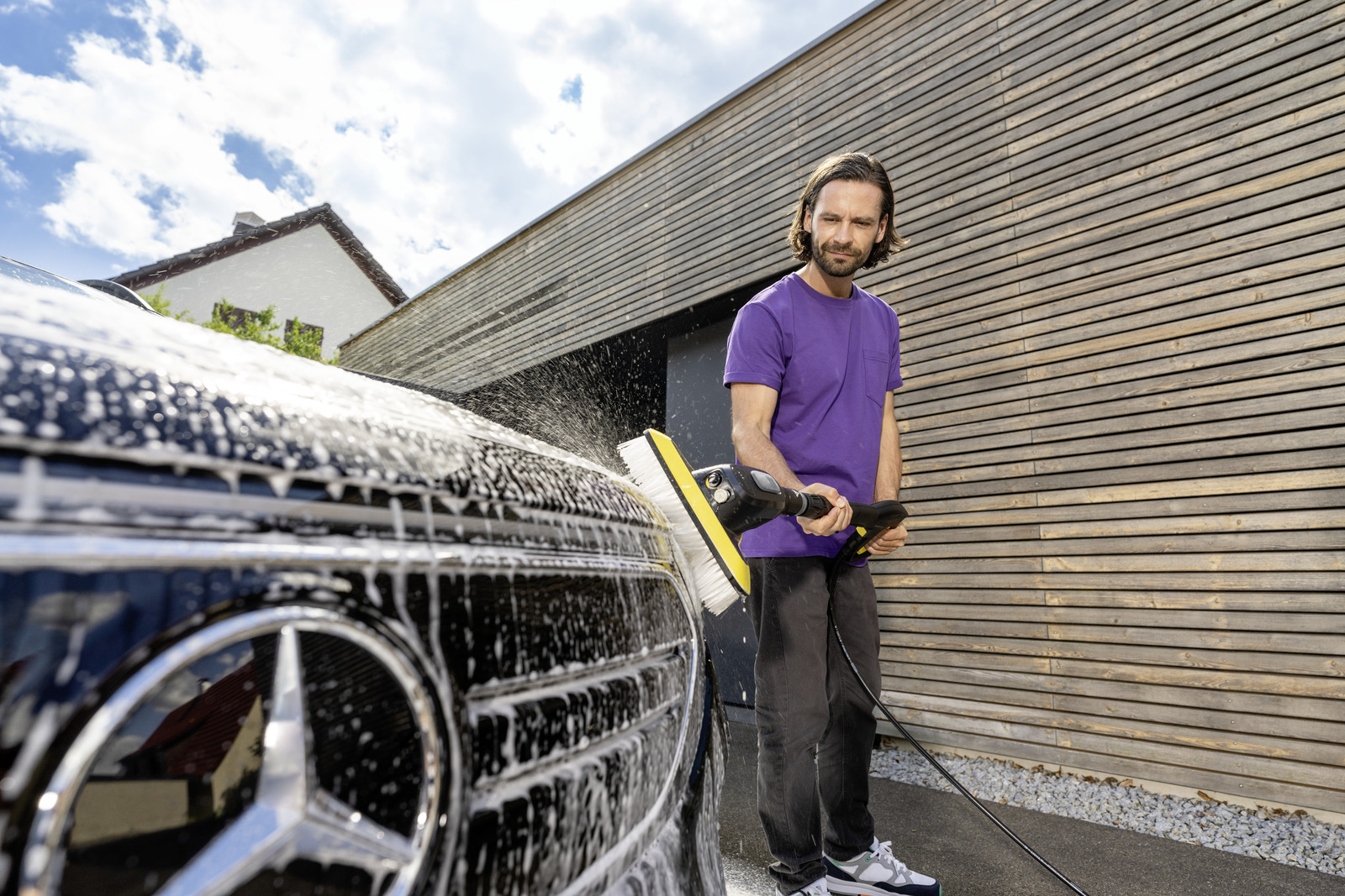 Mann wäscht Auto mit Bürste und Wasserstrahl vor Holzwand.