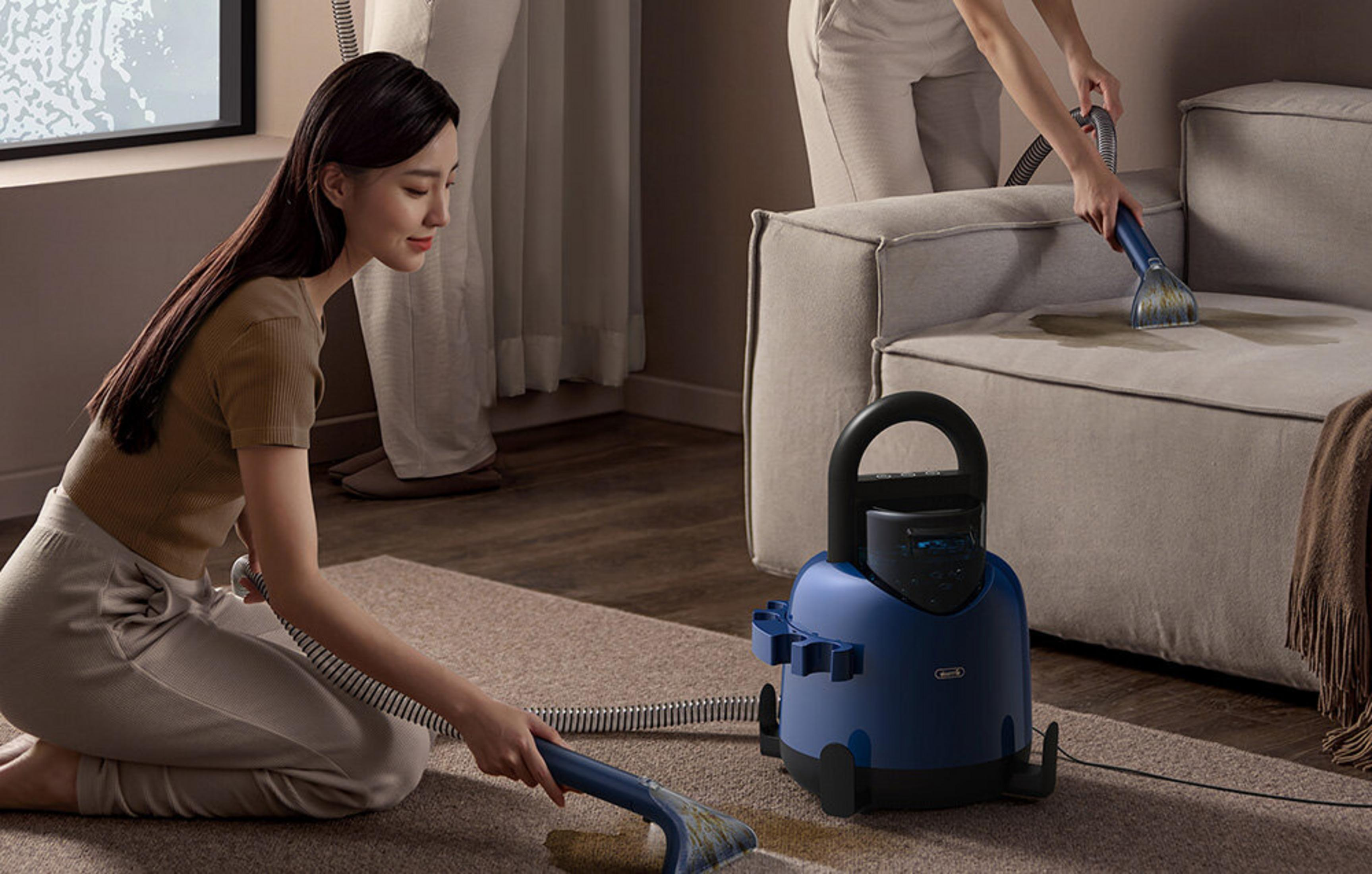 Una donna in ginocchio su un tappeto pulisce una macchia sul divano con un apparecchio blu.