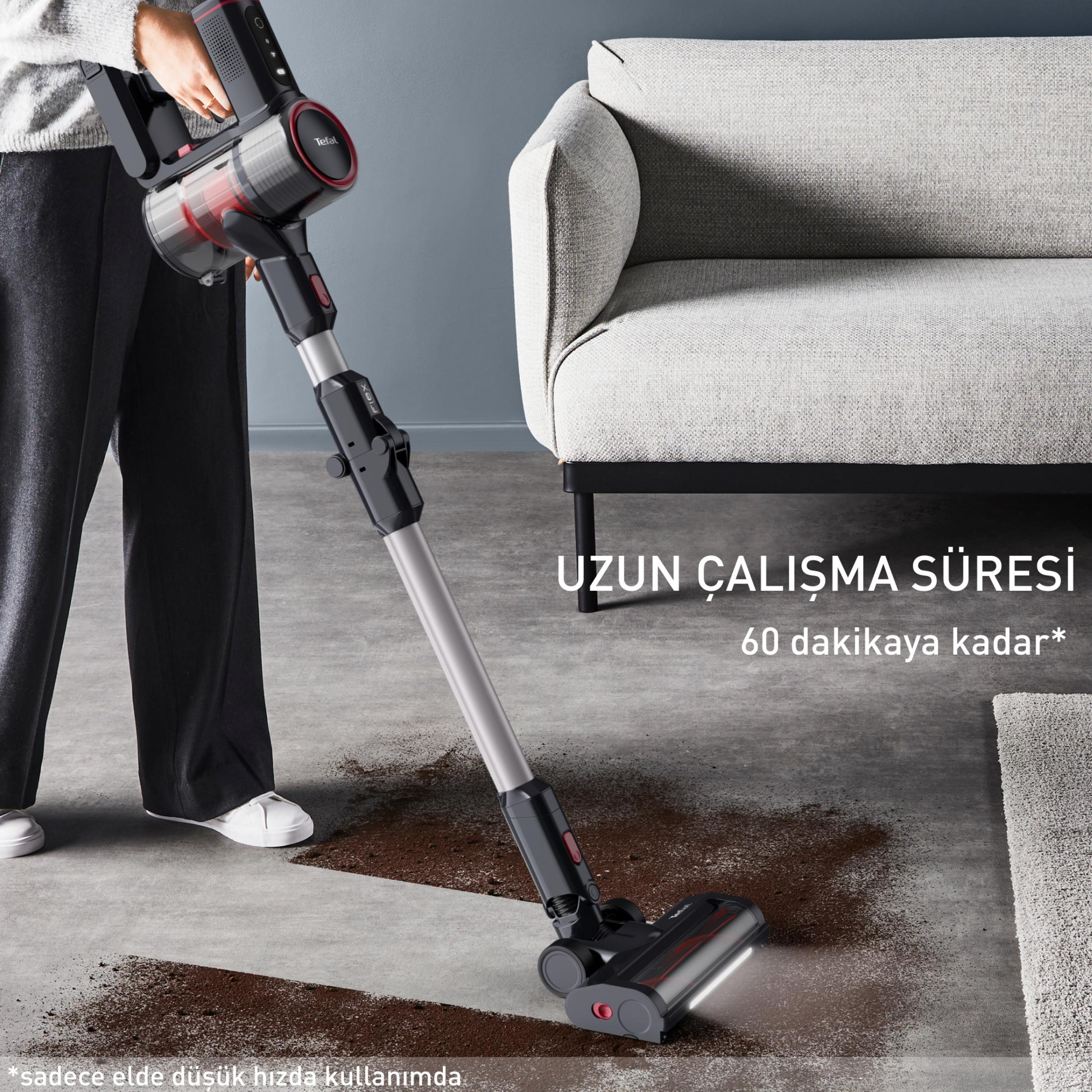 Kişi, siyah elektrikli süpürge ile gri zemindeki kahverengi kiri süpürüyor. Arkada kanepe.