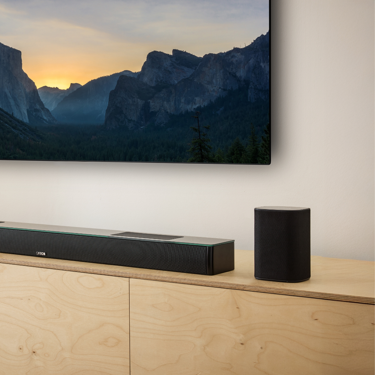 Schwarze Soundbar und zylindrischer Lautsprecher auf hellem Holzmöbel vor TV mit Berglandschaft.